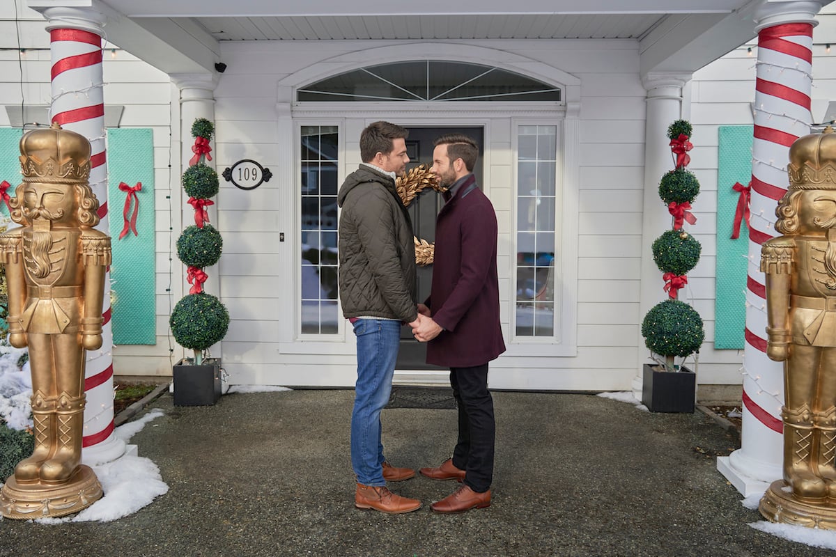 Brandon and Jake holding hands and facing each other in front of a house decorated for Christmas in 'The Christmas House 2: Deck Those Halls'