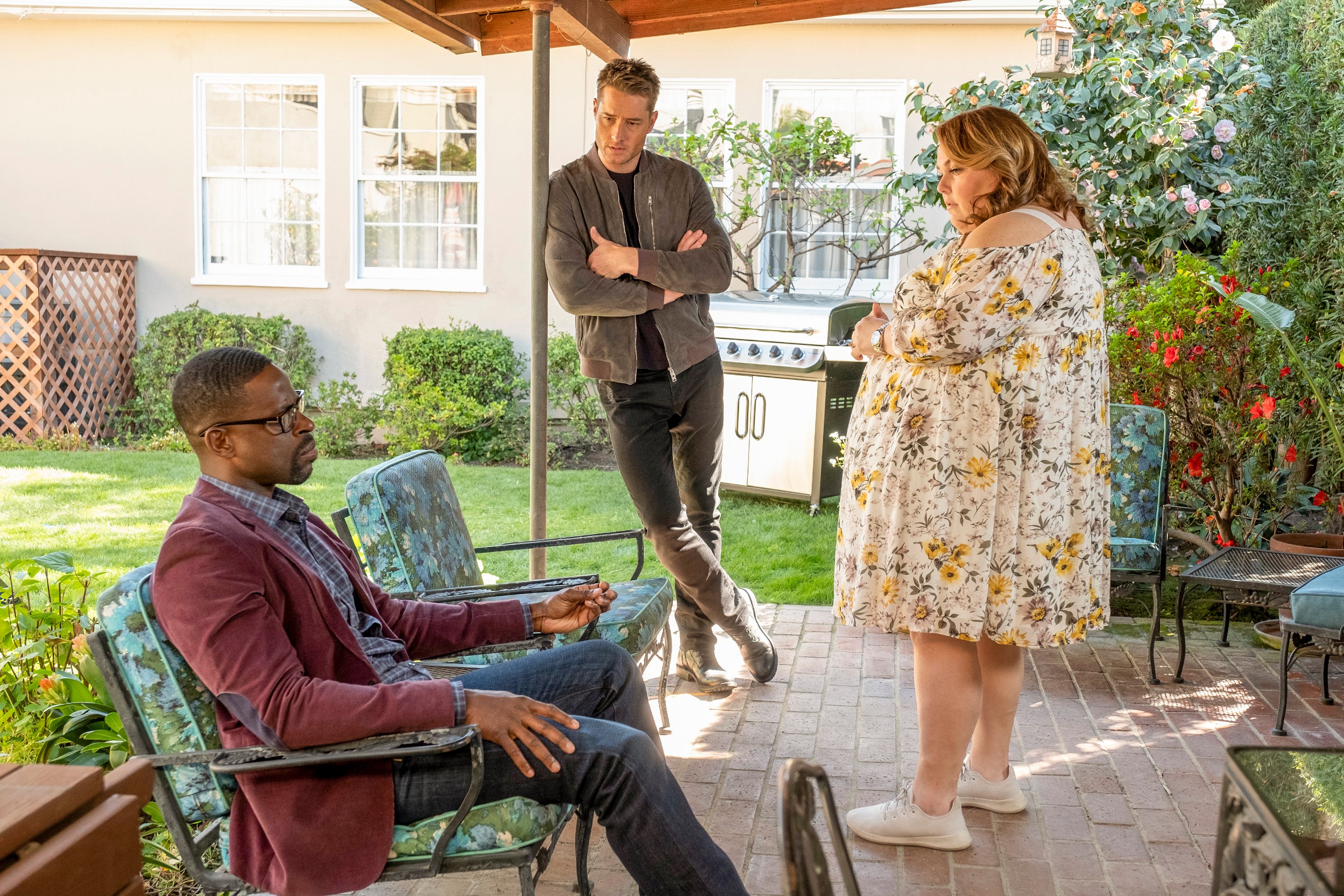 'This Is Us' actors Sterling K. Brown, Justin Hartley, and Chrissy Metz, in character as Randall, Kevin, and Kate in the show, gather in a backyard. Randall, wearing a maroon blazer over a blue plaid shirt and jeans, sits in a lawn chair. Kevin, wearing a gray jacket over a black shirt and light black plants, leans against a pole. Kate stands in front of them wearing a yellow and white floral dress.