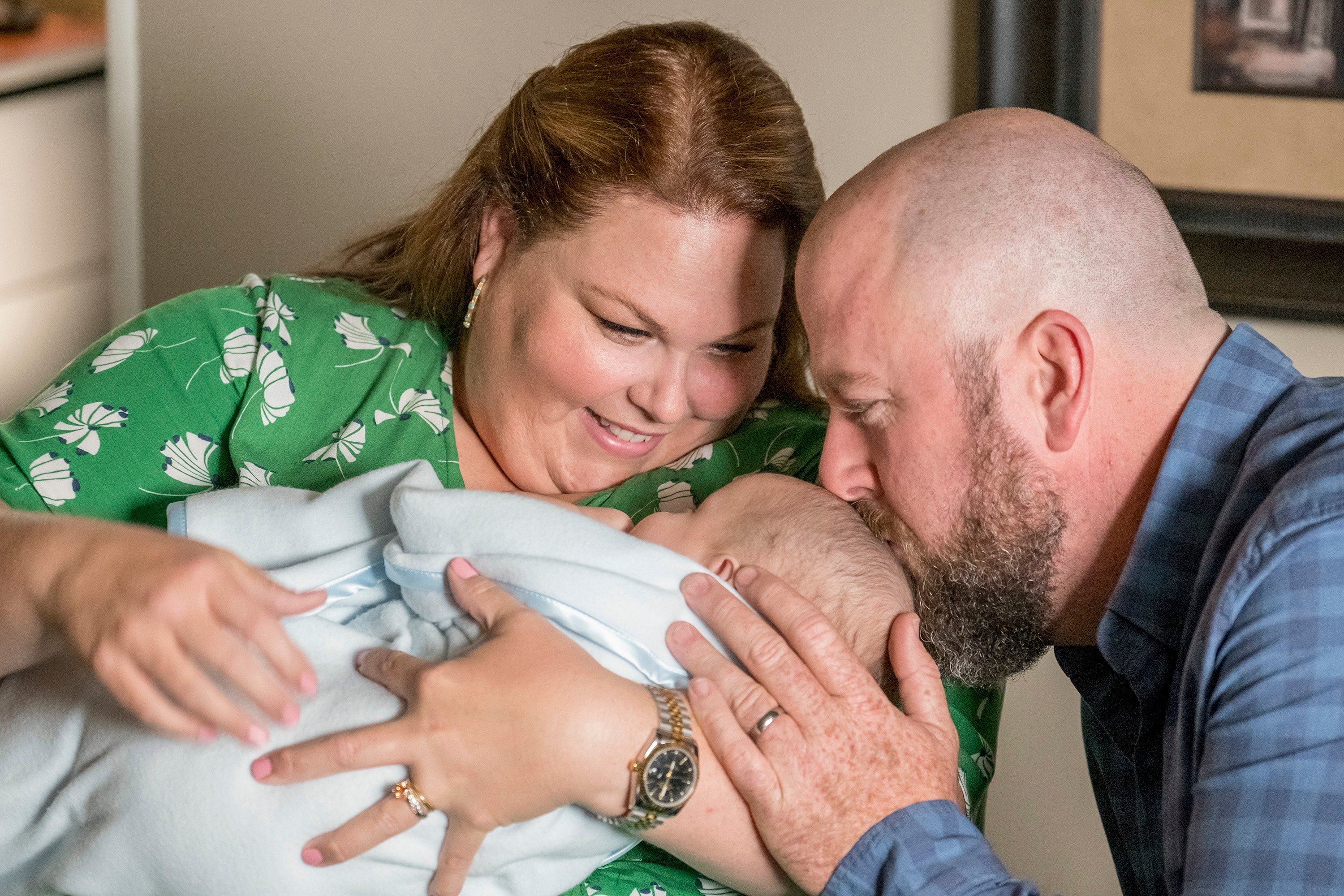 'This Is Us' Season 6 actors Chrissy Metz and Chris Sullivan, in character as Kate and Toby, hold their baby, Jack. Kate wears a green dress with white flowers. Toby wears a blue plaid shirt and kisses Jack's head.