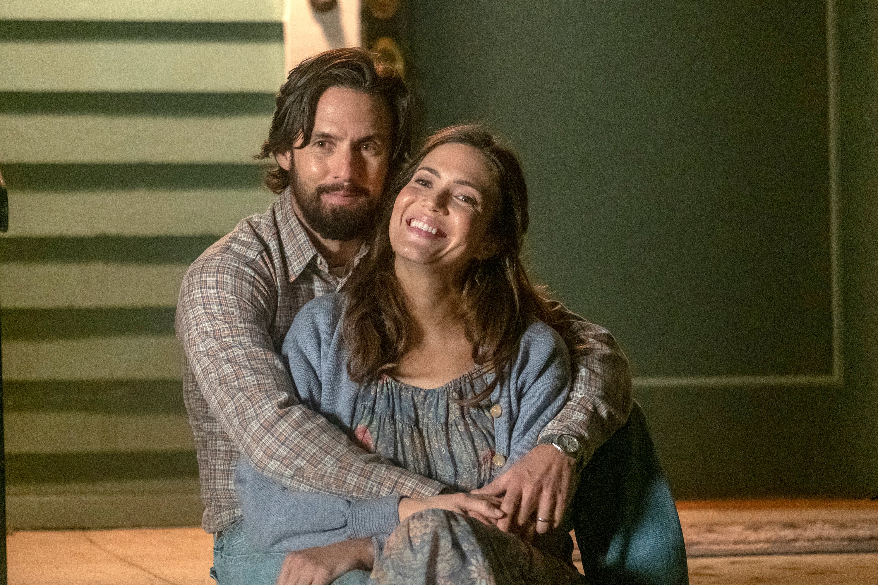 'This Is Us' Season 6 stars Milo Ventimiglia and Mandy Moore, in character as Jack and Rebecca Pearson, sit on their porch with Jack sitting behind Rebecca and his arms around her. Jack wears a light brown and white plaid long-sleeved button-up shirt and jeans. Rebecca wears a gray floral dress and a light blue cardigan.  Both characters will be a part of the ending in 'This Is Us' Season 6.