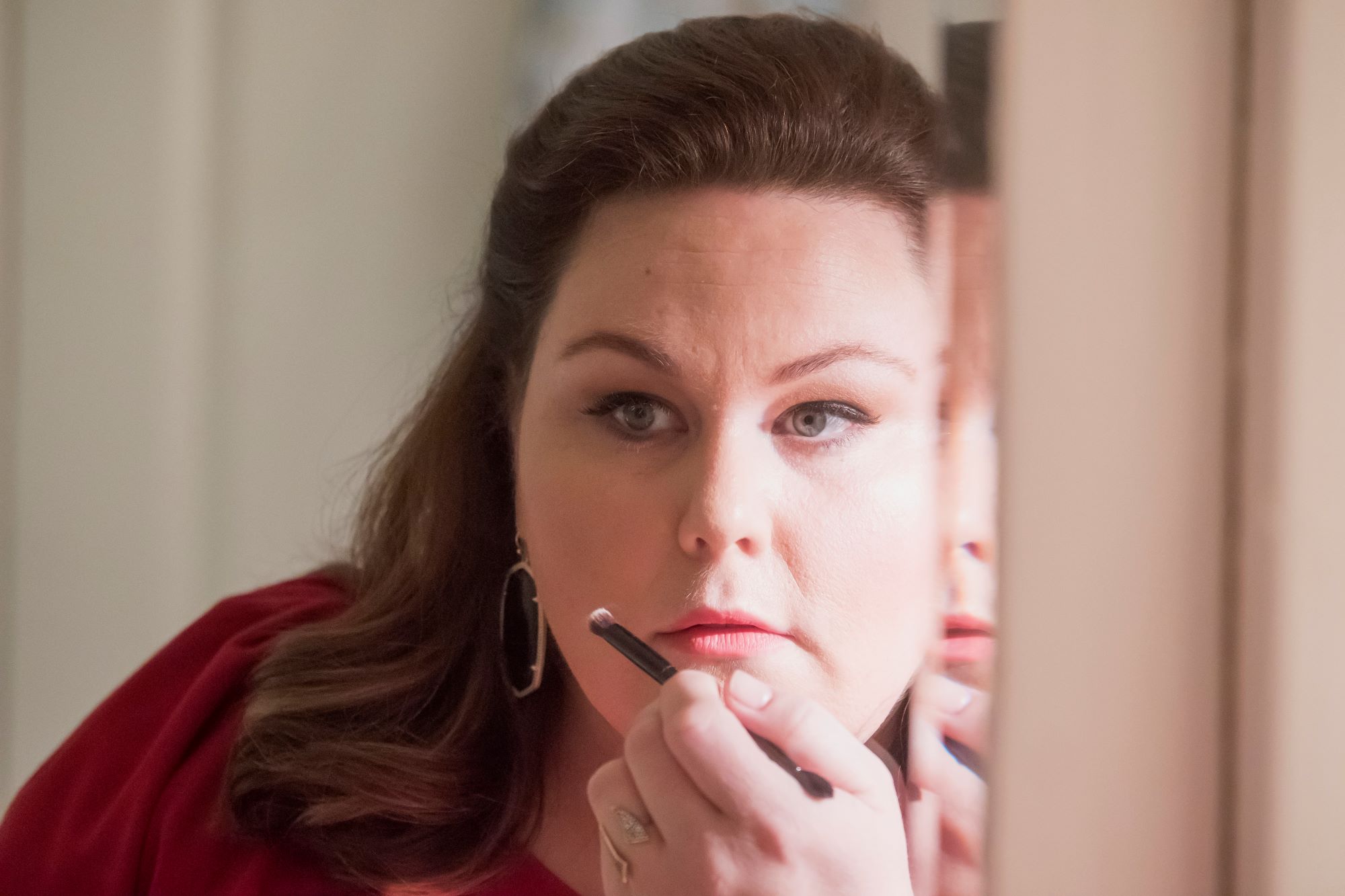'This Is Us' Season 6 star Chrissy Metz, in character as Kate Pearson, wears a red shirt and applies makeup to her face with a small brush as she looks in the mirror.