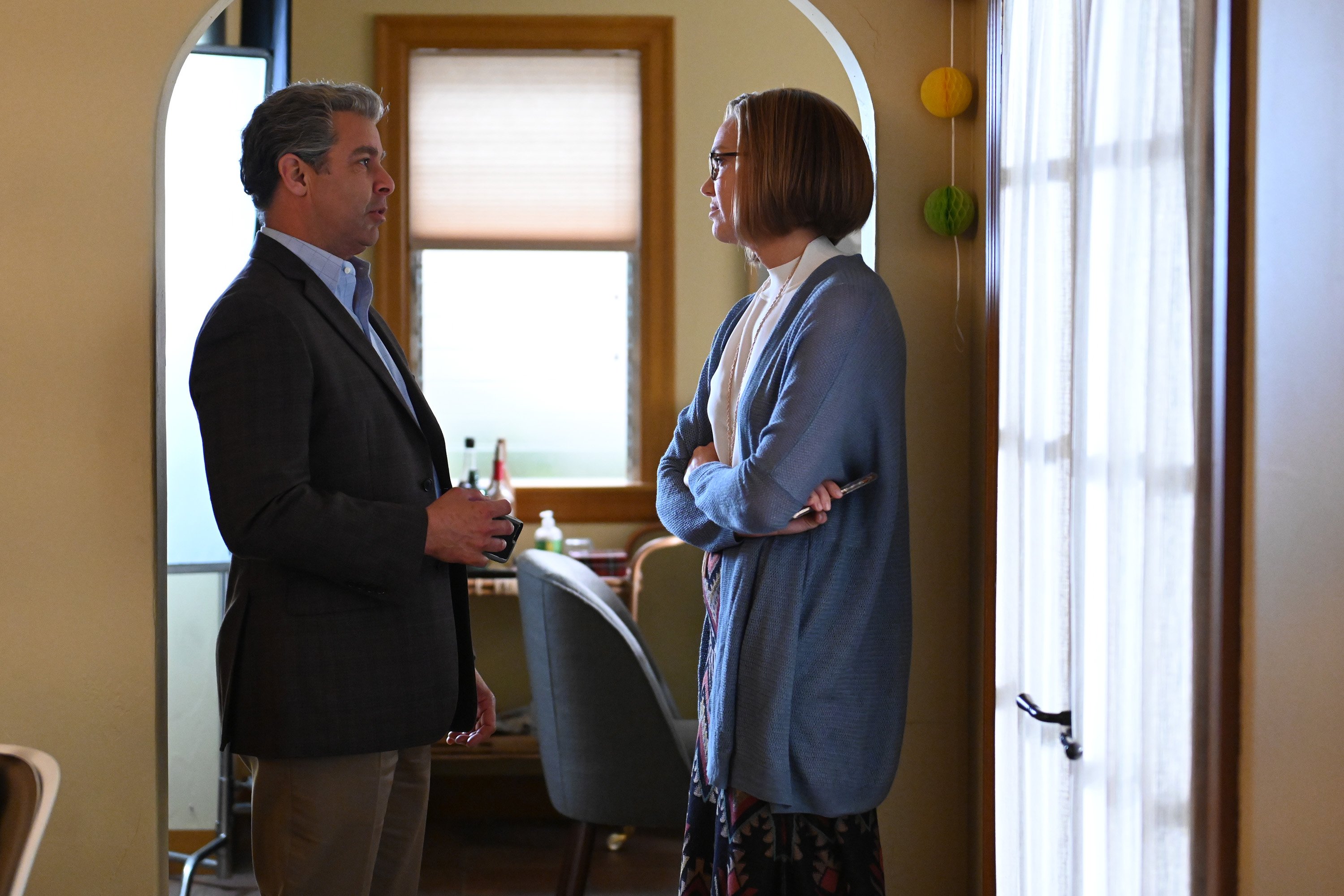 'This Is Us' Season 6 stars Jon Huertas and Mandy Moore, in character as Miguel and Rebecca, face one another. Miguel wears a dark gray suit jacket over a blue button-up shirt and tan pants. Rebecca wears a blue cardigan over a white shirt and a floral skirt.