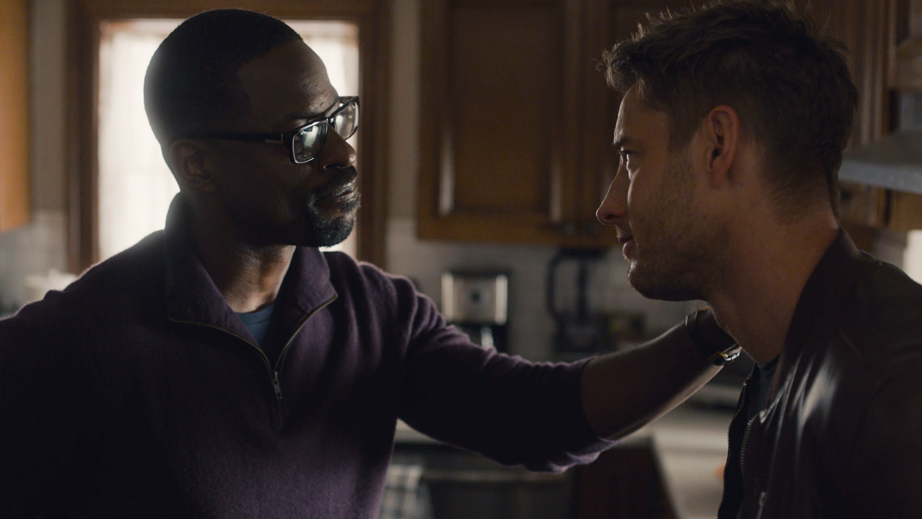 'This Is Us' Season 6 stars Sterling K. Brown and Justin Hartley, in character as Randall and Kevin, talk. Randall, wearing a purple sweater, puts his hand on Kevin's shoulder, who is wearing a dark brown jacket.