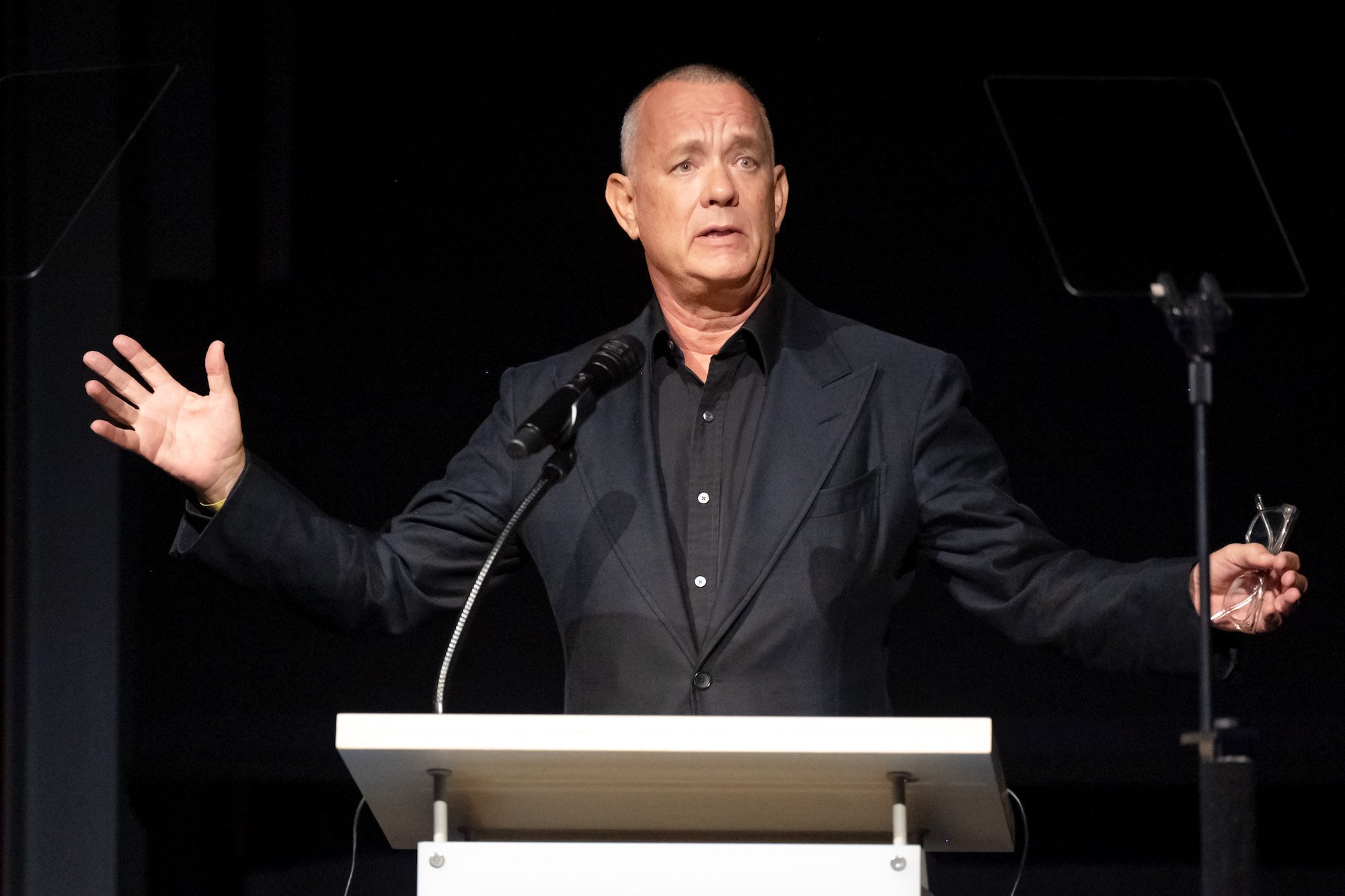 Tom Hanks opens his arms and holds his glasses standing at a podium