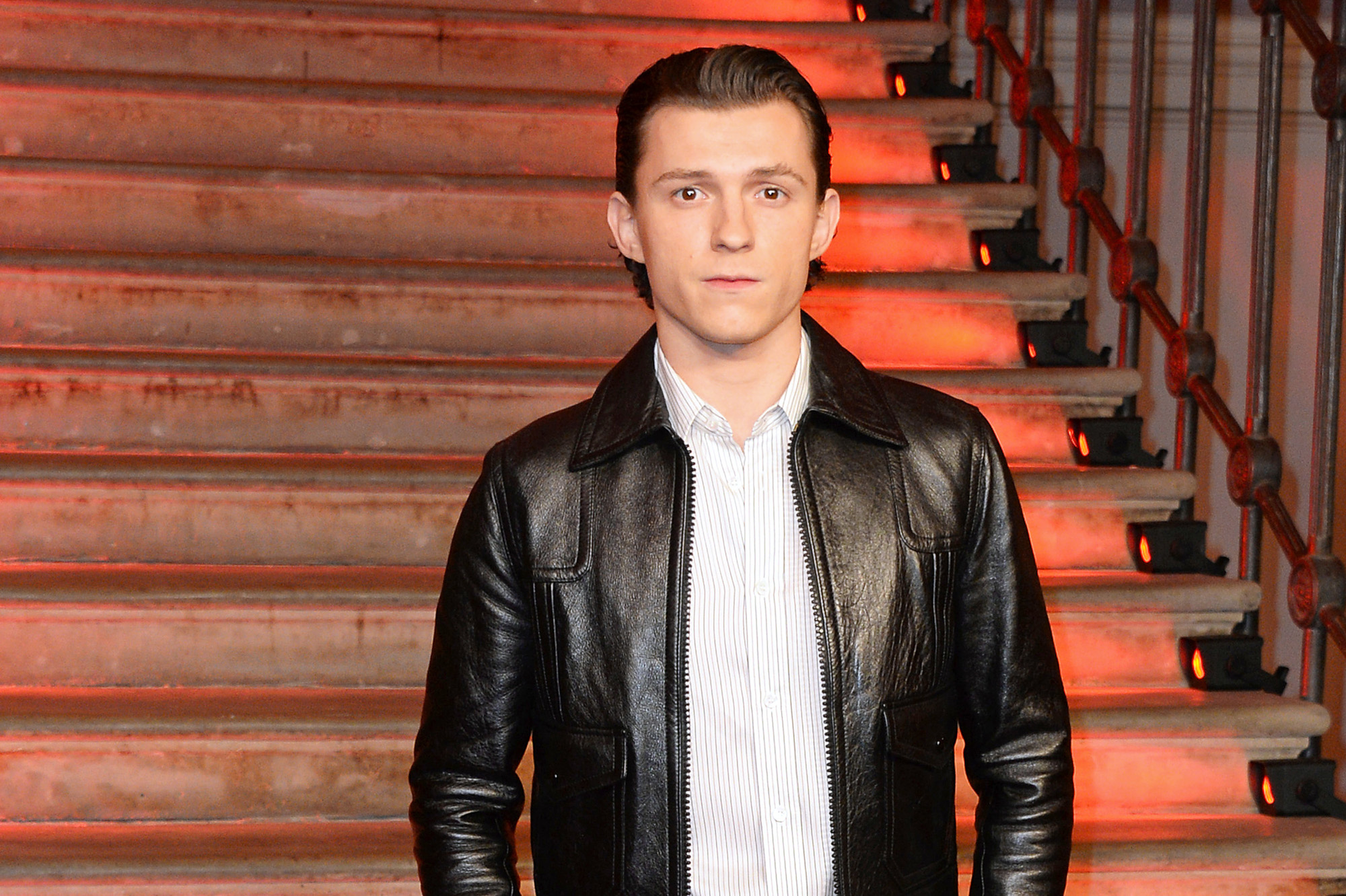 Spider-Man star Tom Holland wearing a black leather jacket and white button-up shirt. His hair is slicked back, and he's looking at the camera.