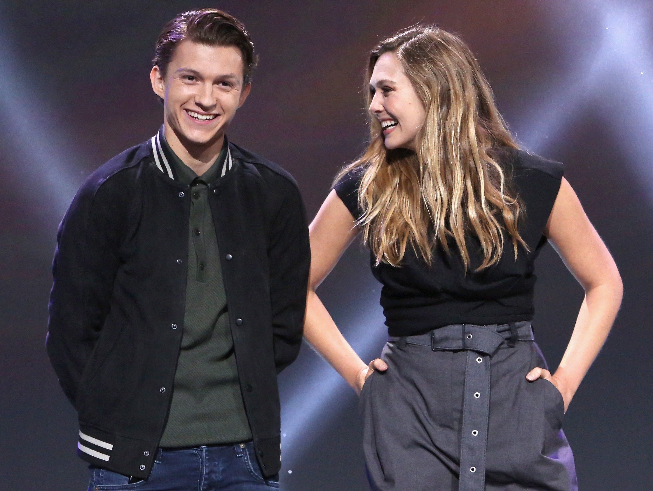 'Spider-Man' actor Tom Holland and Marvel co-star Elizabeth Olsen. They're standing on-stage. He's looking at the camera, and she's looking at him and laughing.