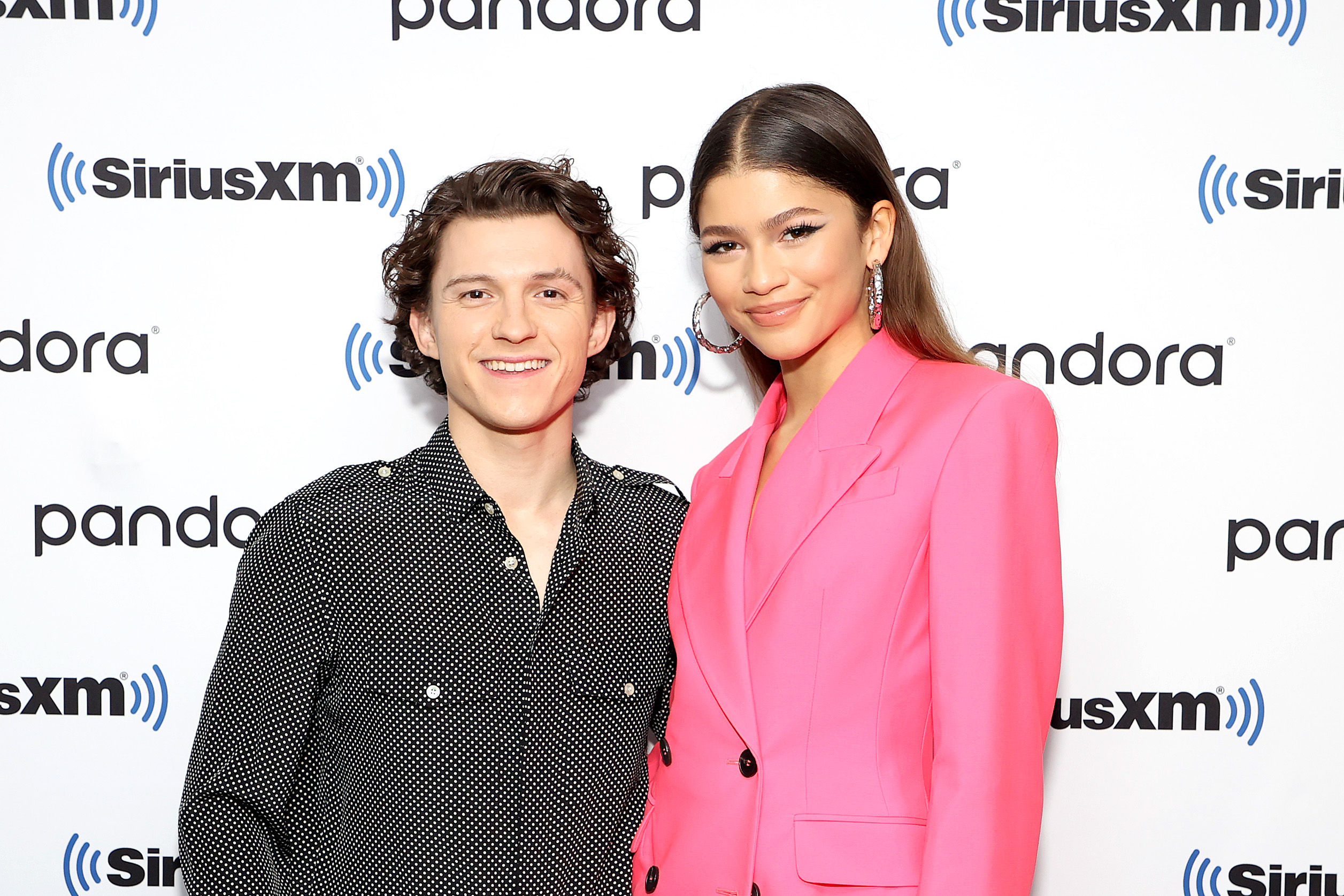 'Spider-Man: No Way Home' stars Tom Holland and Zendaya pose for a picture toether. Holland wears a black button-up long-sleeved shirt with white polka dots. Zendaya wears a bright pink suit.