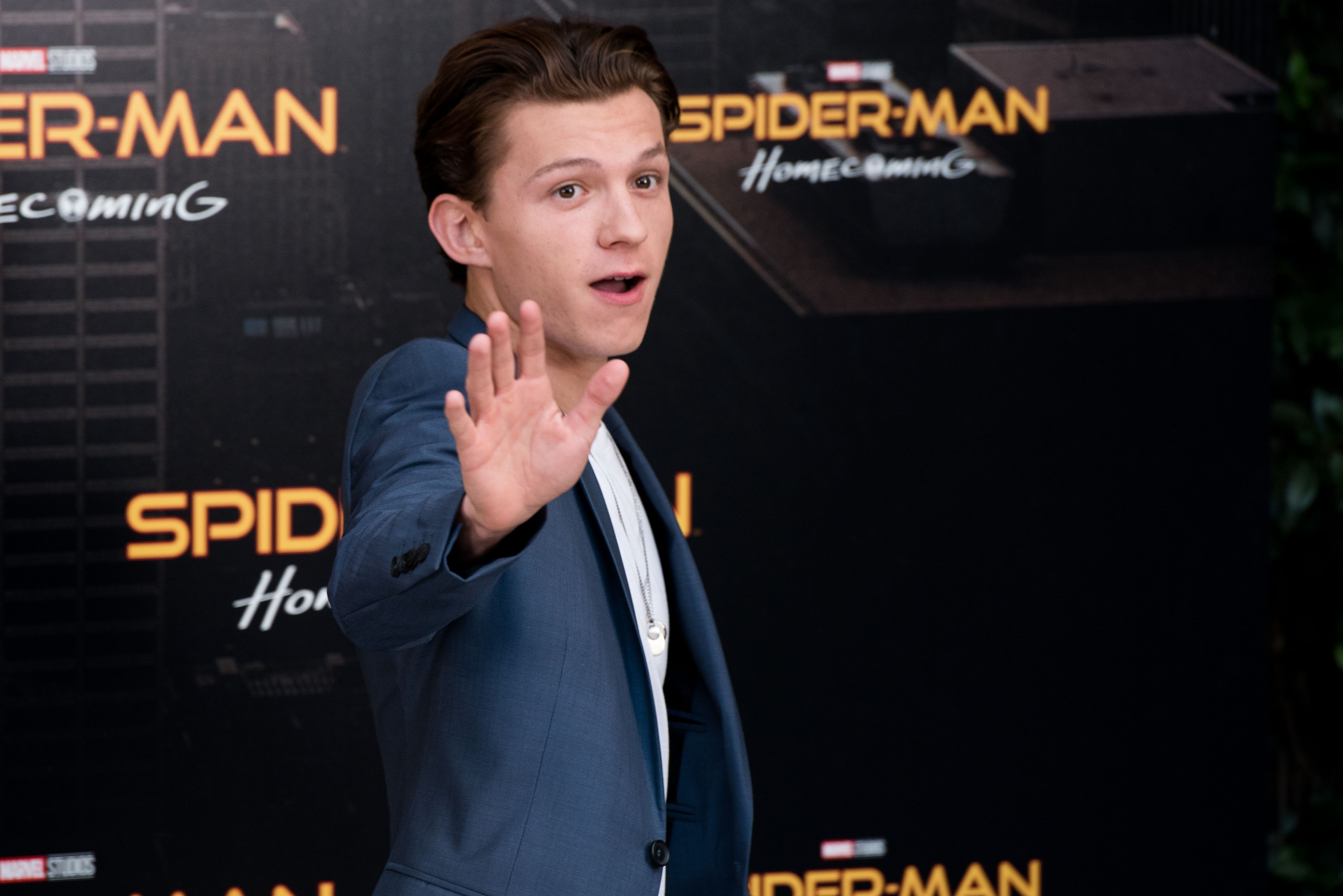 'Spider-Man: Homecoming' star Tom Holland waves to the camera, wearing a blue suit jacket over a white shirt.