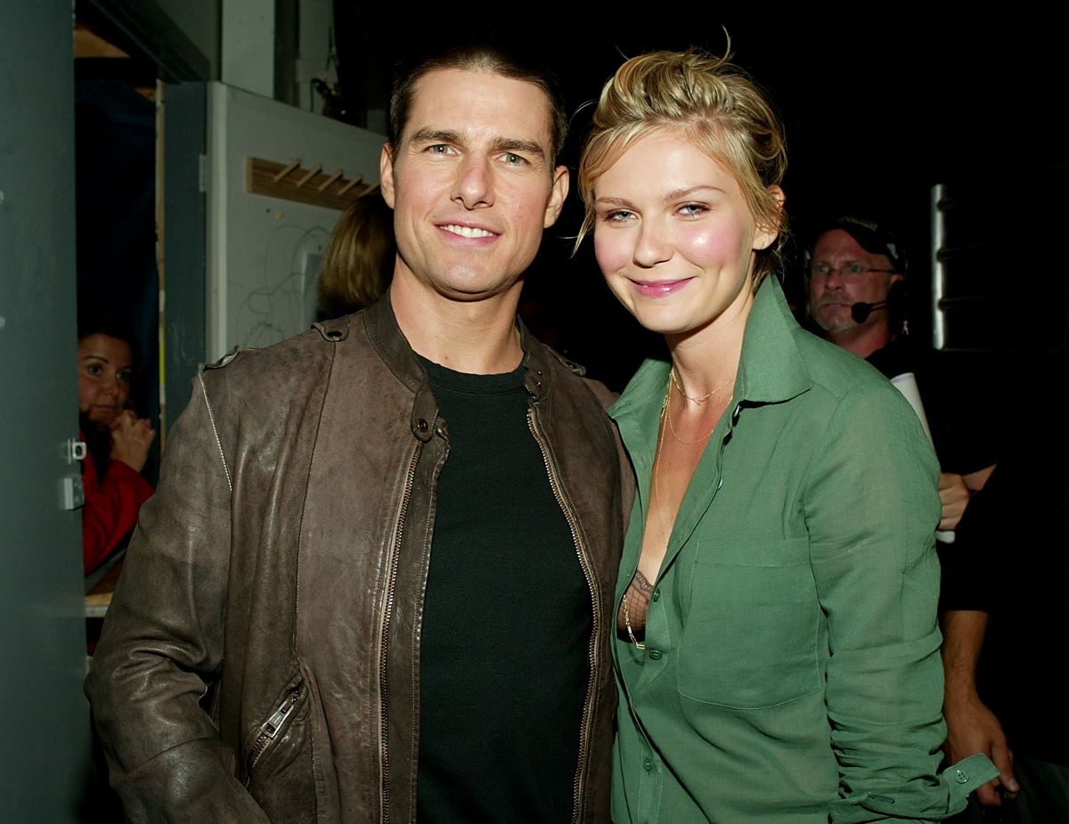 Tom Cruise in a brown jacket and black shirt, stands next to Kirsten Dunst in a green jacket