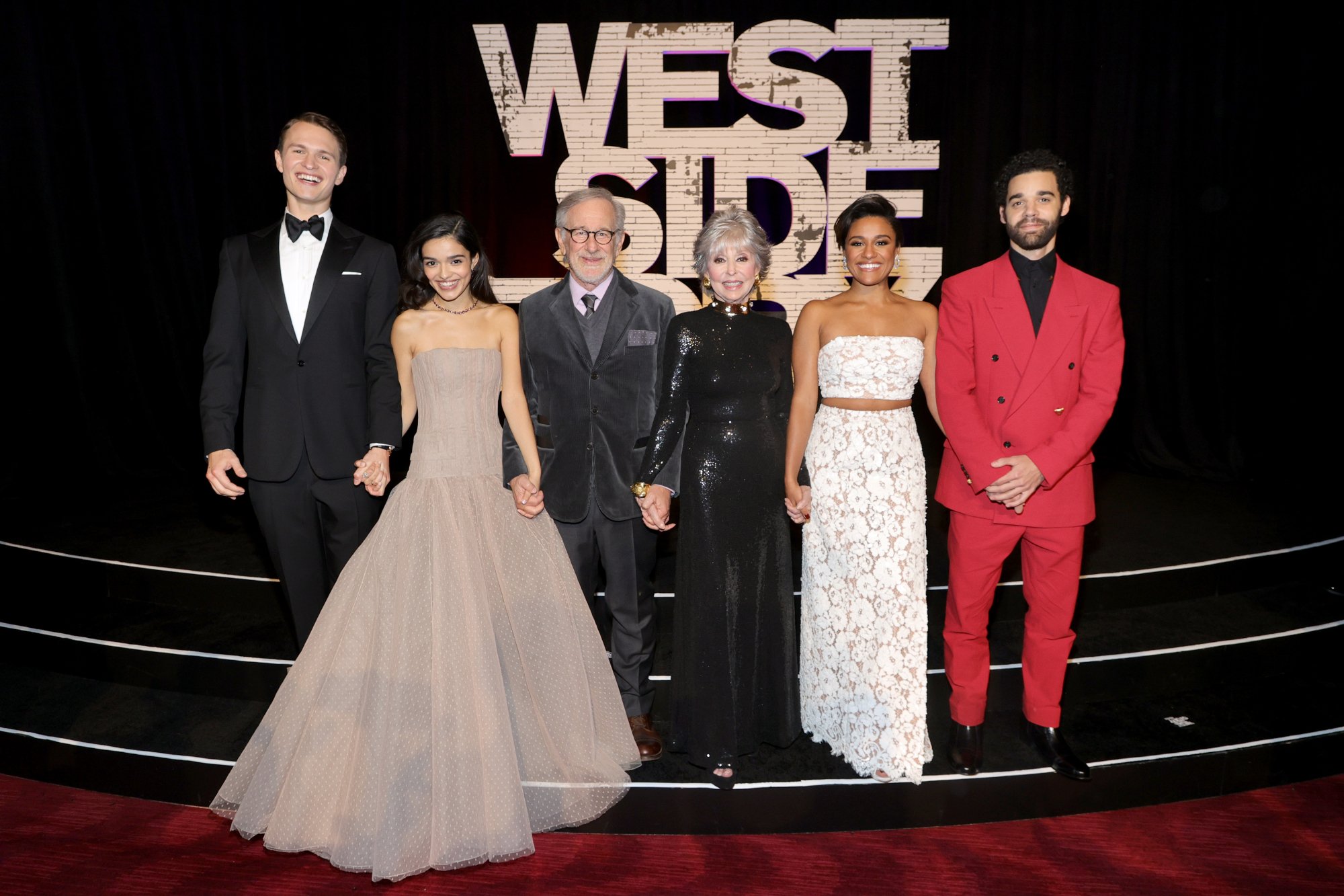 'West Side Story' Ansel Elgort, Rachel Zegler, Steven Spielberg, Rita Moreno, Ariana DeBose, and David Alvarez holding hands in front of the movie title logo