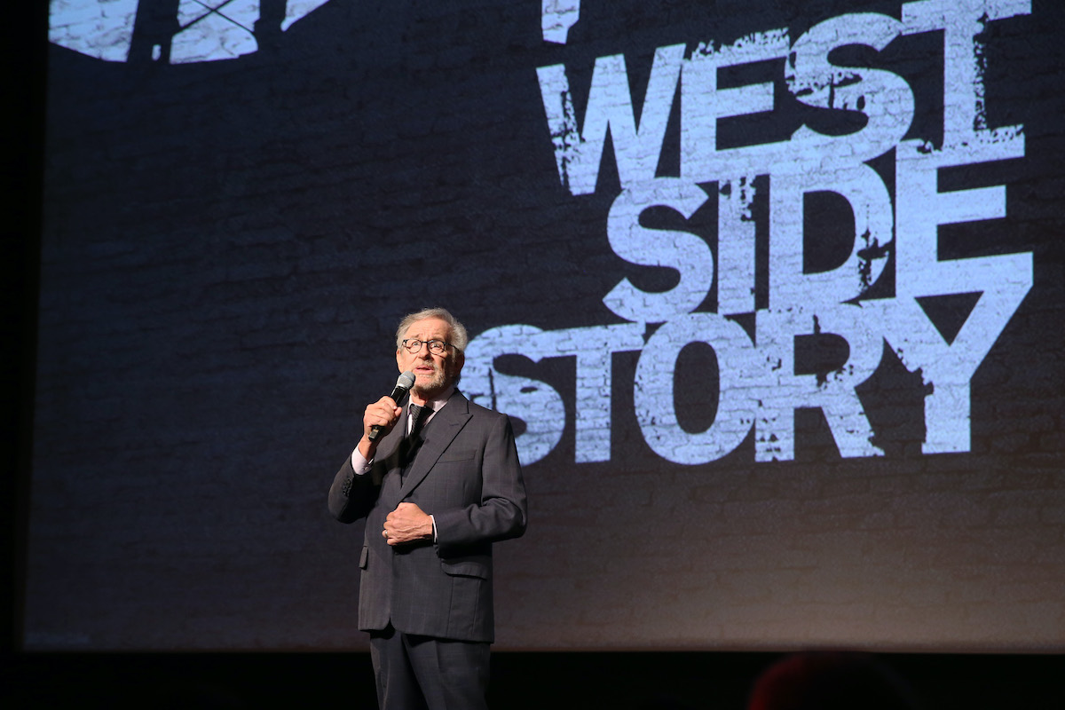 Steven Spielberg wears a suit and speaks onstage in front of the logo of his ‘West Side Story’