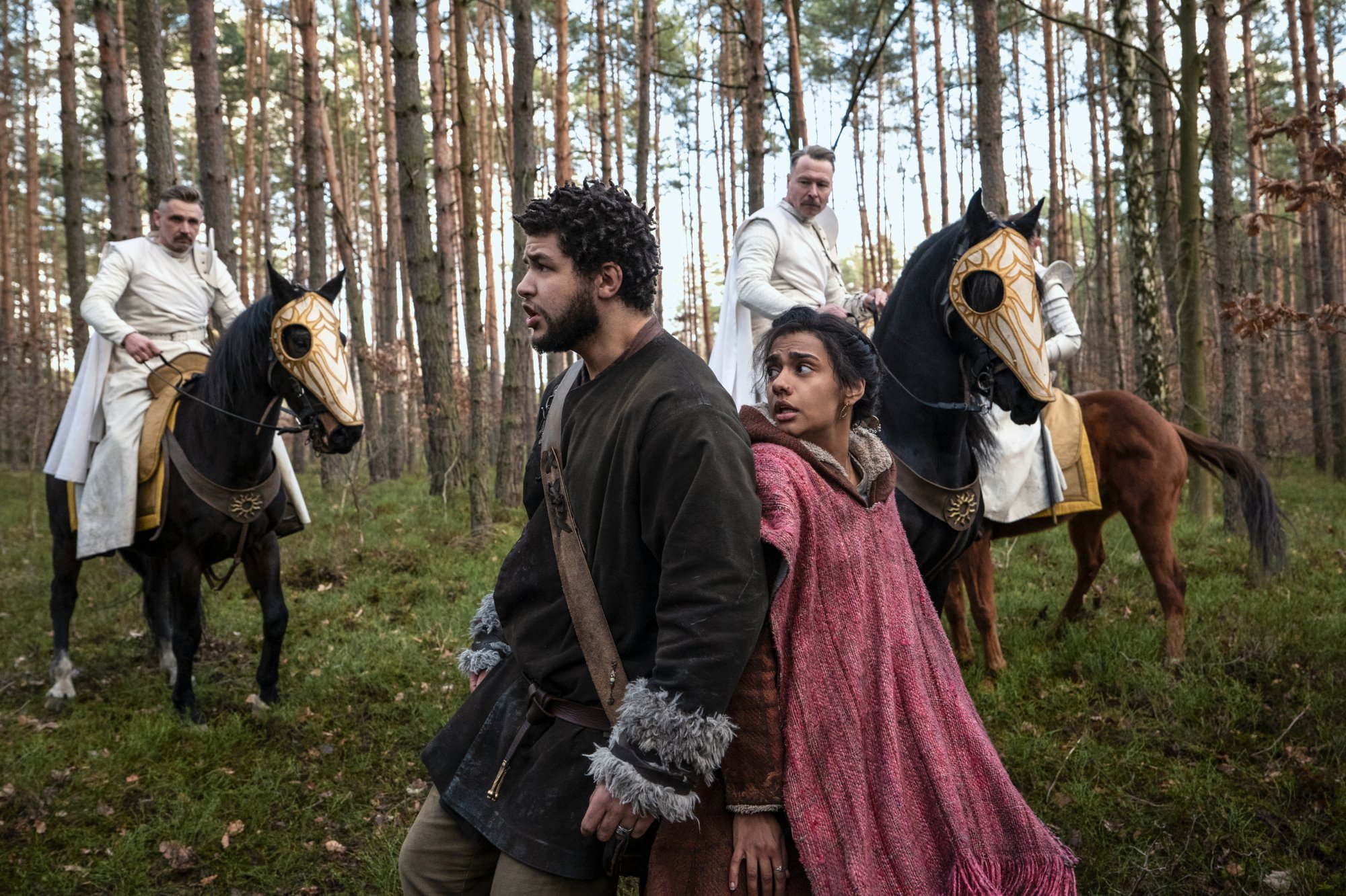 Marcus Rutherford and Madeleine Madden in 'The Wheel of Time' Episode 5. They're standing back to back and being surrounded by men wearing white and riding on horseback.