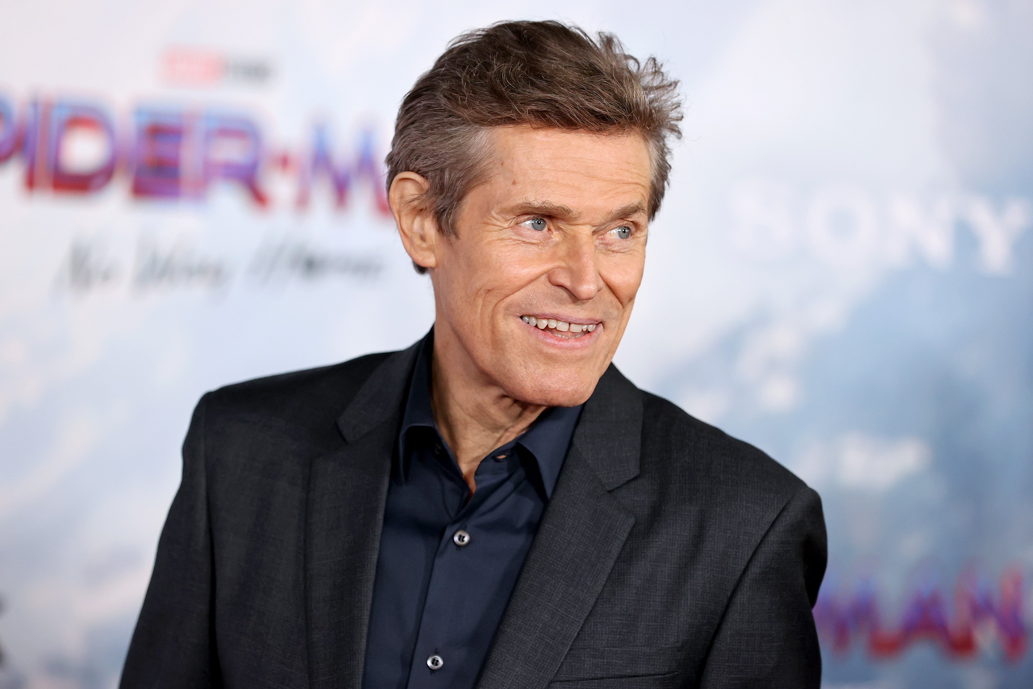 Green Goblin actor Willem Dafoe at the premiere for 'Spider-Man: No Way Home.' He's wearing a black suit and smiling at something to the side of the camera.