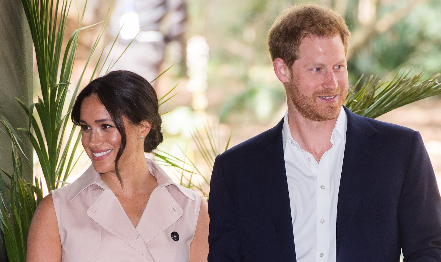 Meghan Markle and Prince Harry smile during their 2019 royal tour in South Africa, with Meghan wearing a tan dress and Harry wearing a white shirt and dark jacket