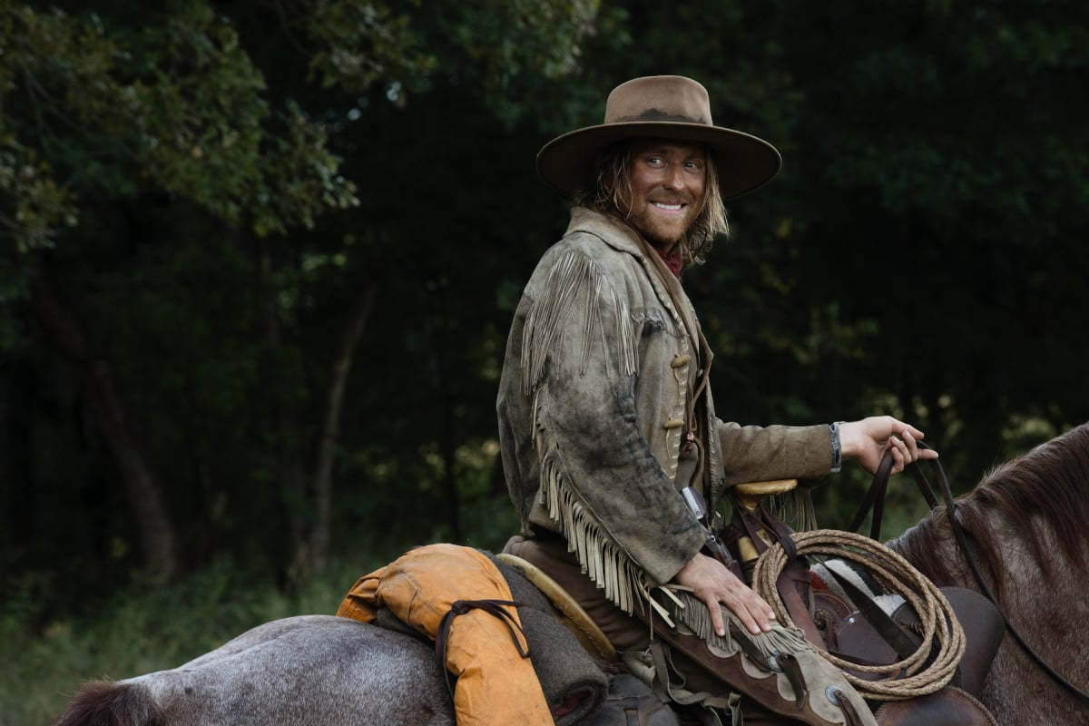 Eric Nelsen as Ennis of the Paramount+ original series 1883. Ennis rides a horse and looks back and smiles.