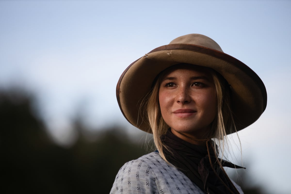 Isabel May as Elsa of the Paramount+ original series 1883. Elsa smiles, wearing a blue dress and brown hat.