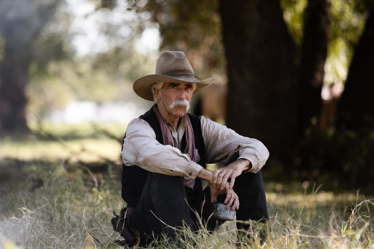 Sam Elliott as Shea of the Paramount+ original series 1883. Shea sits in the grass wearing a cowboy hat.