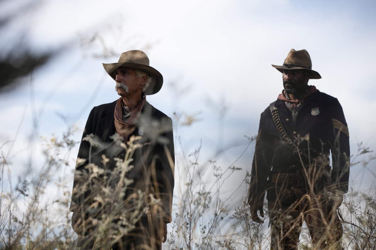 Sam Elliott as Shea and LaMonica Garrett as Thomas of the Paramount+ original series 1883. Thomas and Shea stand side by side behind some weeds.