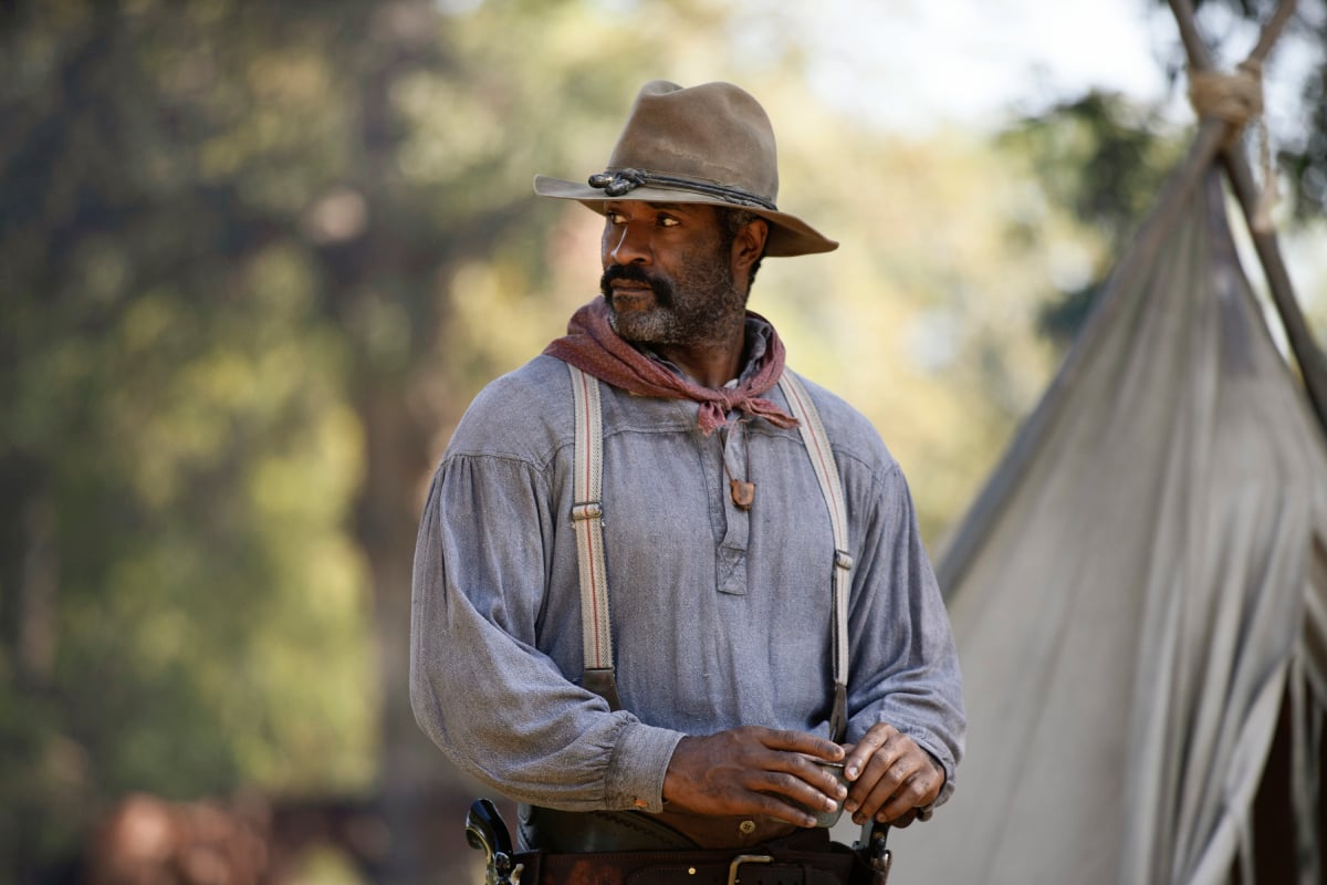 LaMonica Garrett as Thomas of the Paramount+ original series 1883. Thomas wears a long-sleeved shirt, suspenders, and a cowboy and stands in front of a tent.
