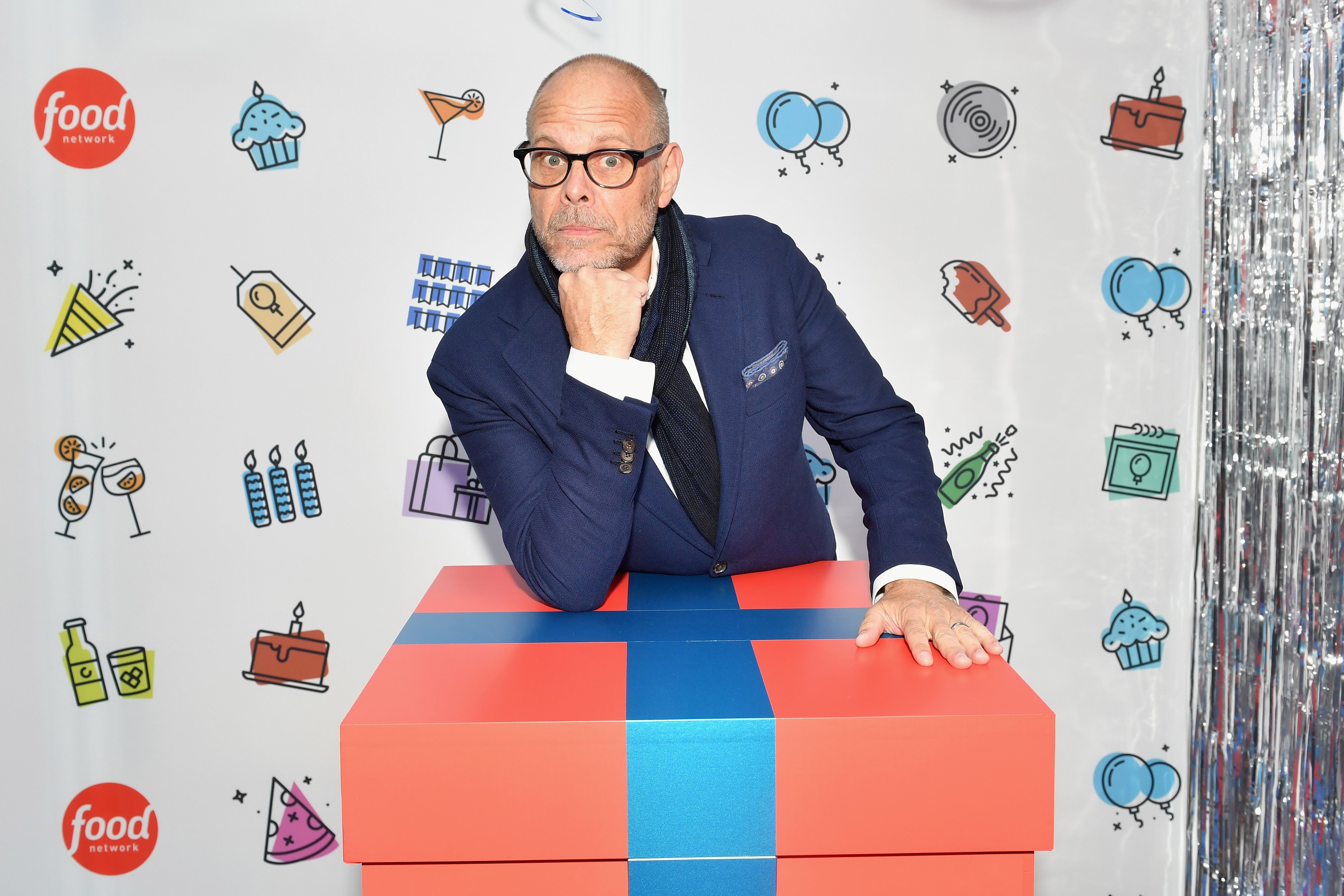 Food Network host Alton Brown poses with hand under chin on a large gift box at a 2018 Food Network event.