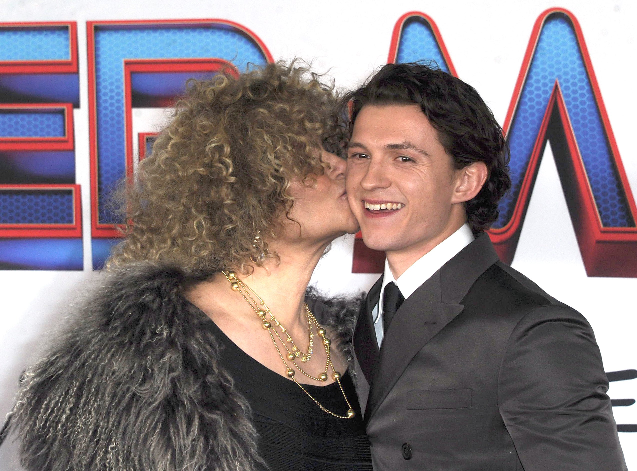Amy Pascal, producer of 'Spider-Man: No Way Home,' kisses actor Tom Holland on the cheek on the red carpet for the premiere. Pascal wars a gray fluffy shawl over a black top and a gold necklace. Holland wears a dark gray suit over a white button-up shirt and black tie. Both Pascal and Holland are pushing for the Oscars to recognize 'Spider-Man: No Way Home.'