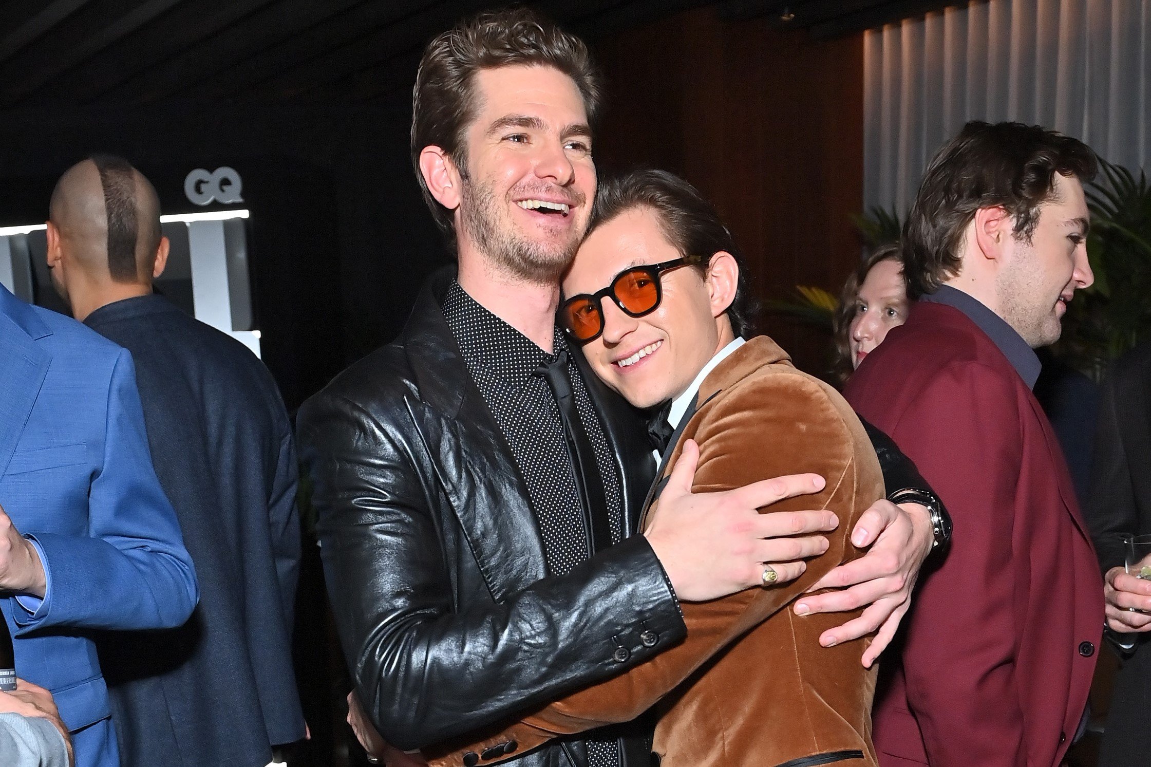 'Spider-Man: No Way Home' stars Andrew Garfield and Tom Holland embrace. Garfield wears a black leather jacket over a black and white polka dotted button-up shirt and black tie. Holland wears a brown velvet blazer over a white button-up shirt and orange-tinted glasses.