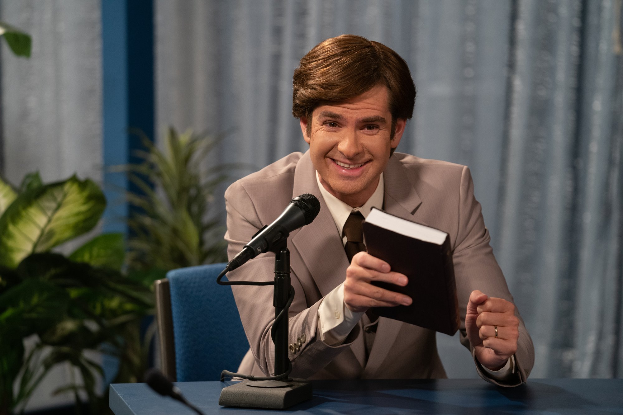 Andrew Garfield as Jim Bakker in 'The Eyes of Tammy Faye' holding a book in front of a microphone