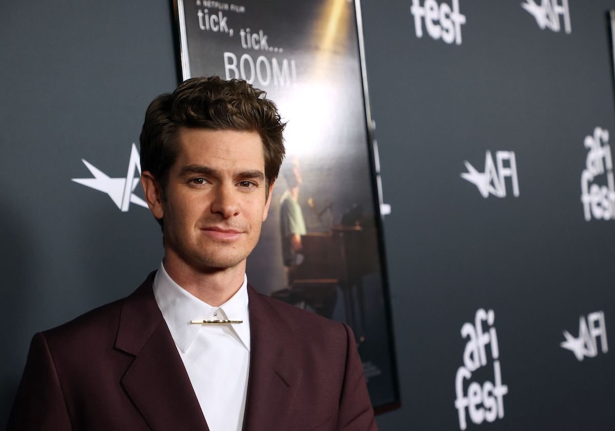 Andrew Garfield wears a suit and poses in front of the ‘Tick, Tick… Boom!’ poster