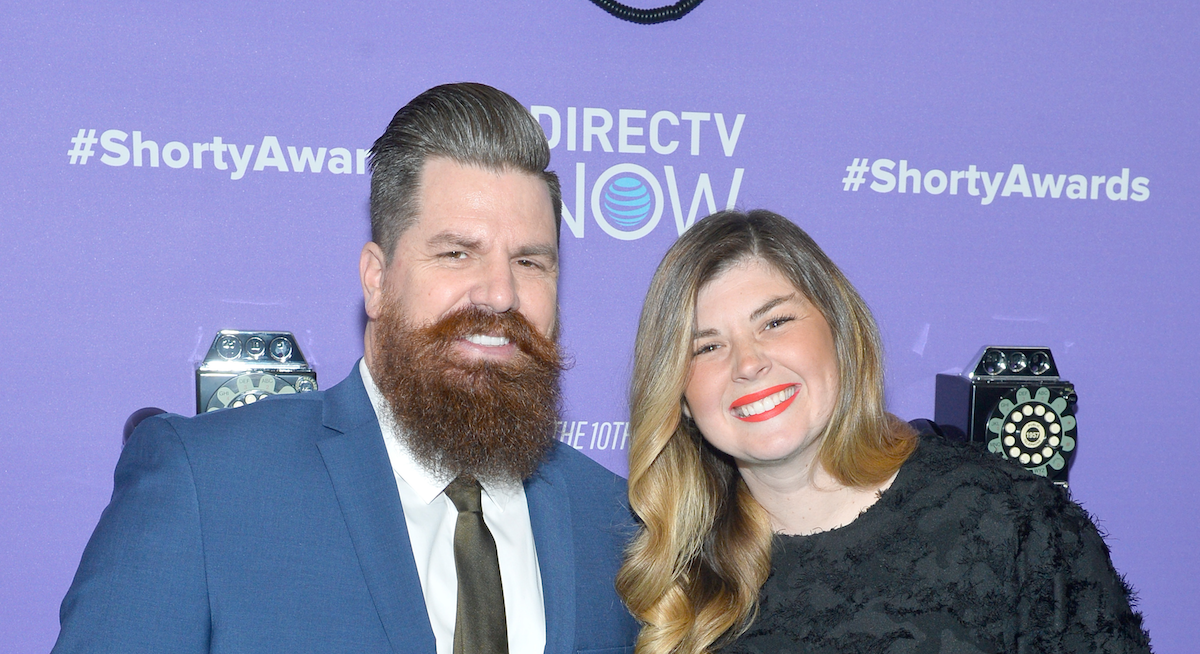 Andy and Candis Meredith of 'Home Work' on the Magnolia Network smile and pose in front of a purple backdrop. Andy Meredith wears a blue suit and Candis Meredith wears a black top.
