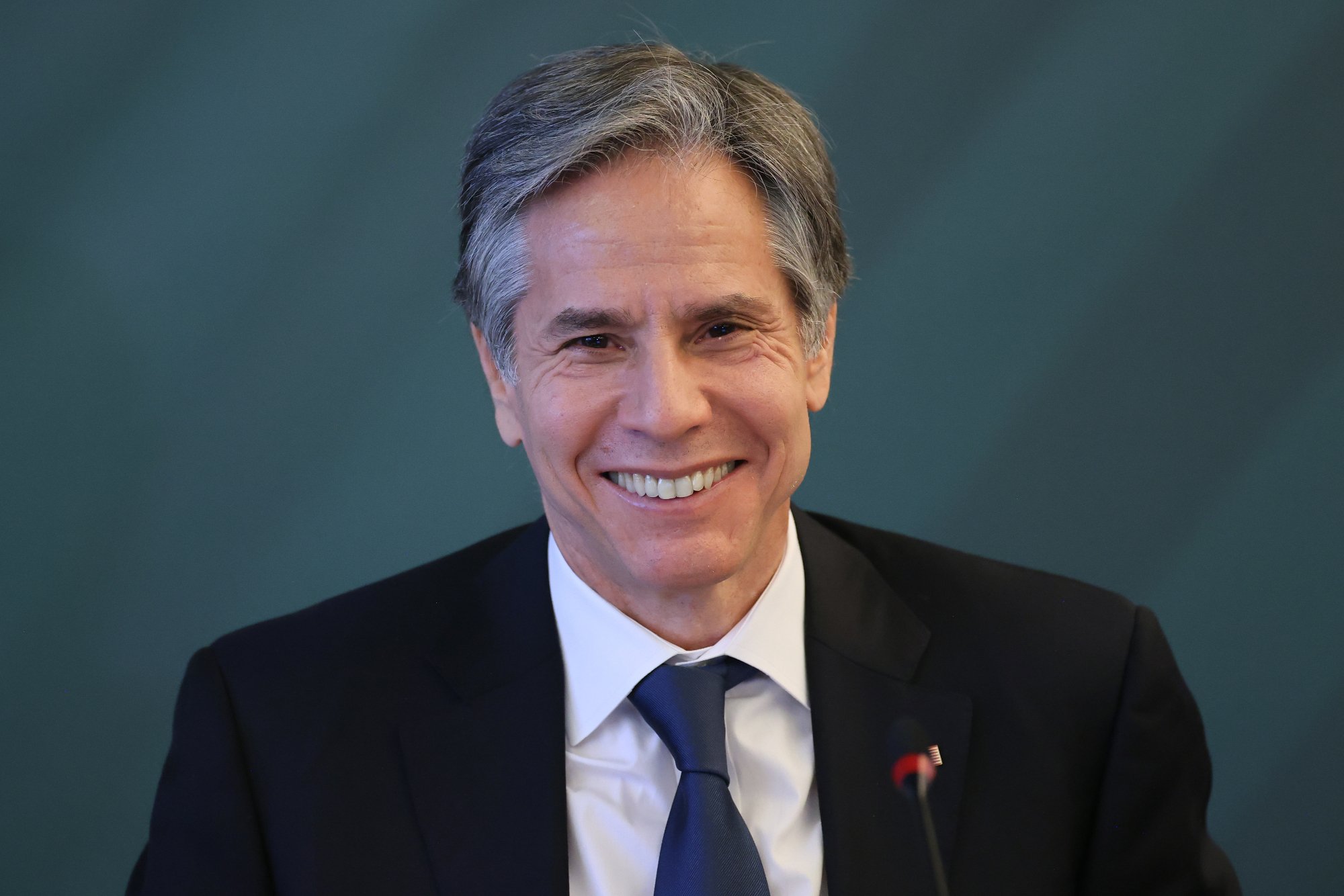 U.S. Secretary of State Antony Blinken. He's wearing a white shirt, black jacket, and blue tie and smiling at the camera. 