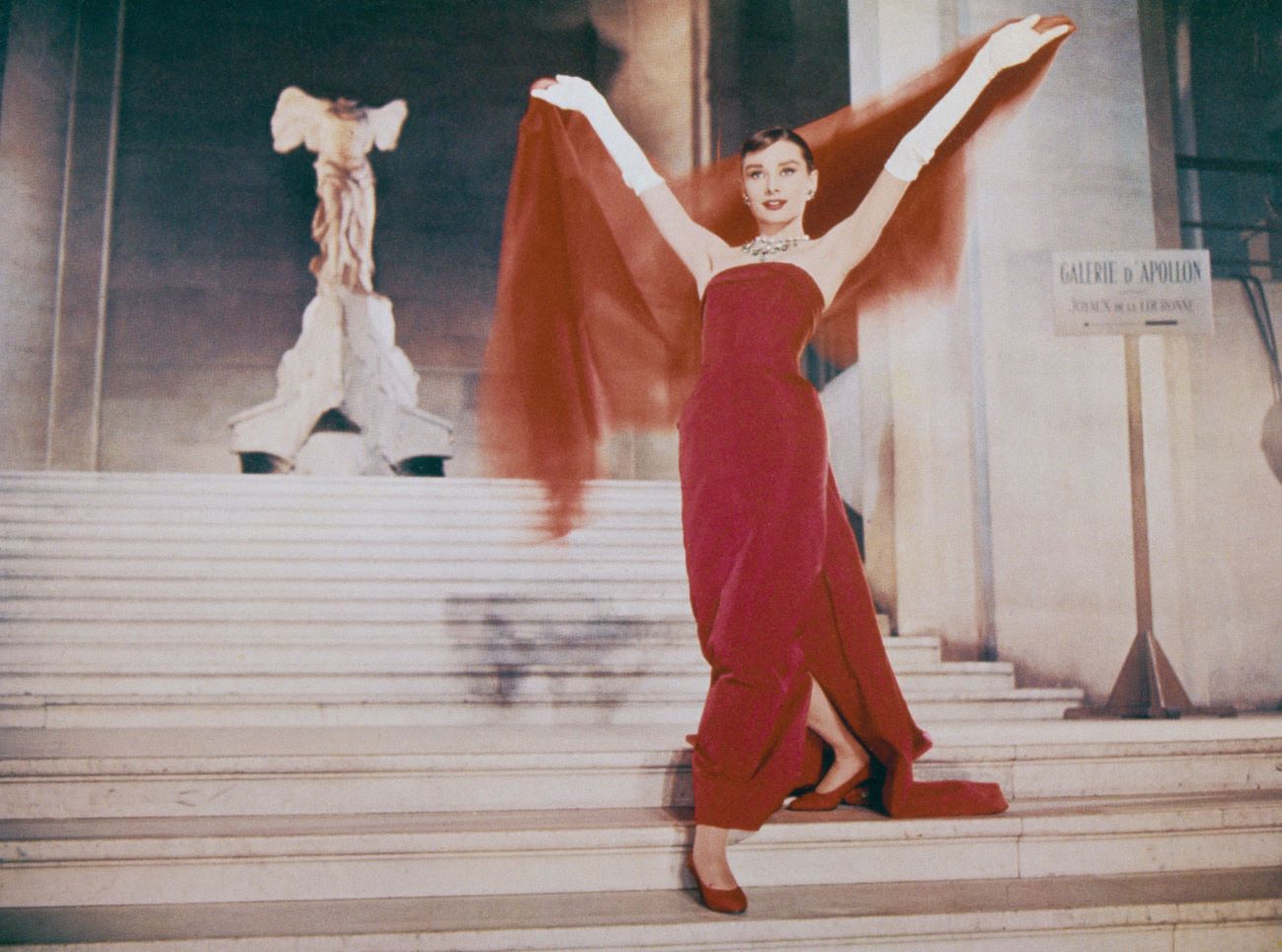 Audrey Hepburn wears a red dress and descends the stairs. She holds a red scarf above her head.