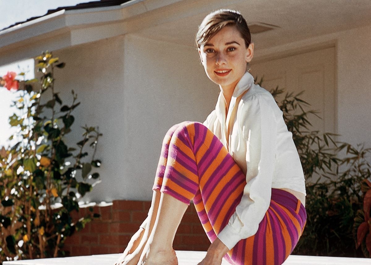 Audrey Hepburn sits with her knees pulled up, wearing a white shirt and colorful, striped pants