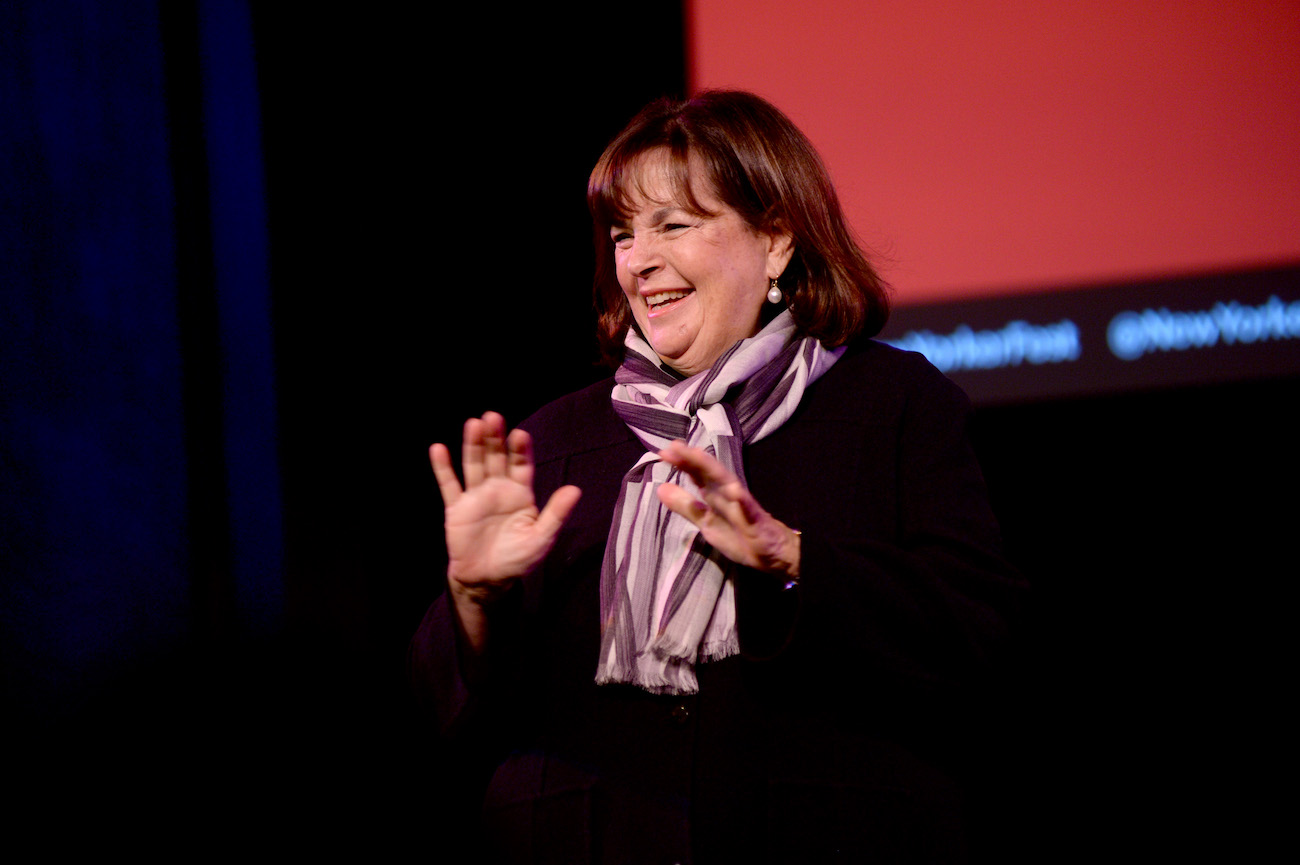 Ina Garten smiles and raises her hands wearing a black top and scarf