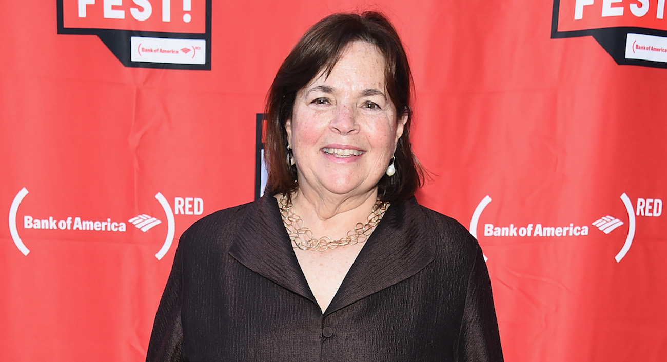 Ina Garten smiles wearing a brown shirt in front of a red background