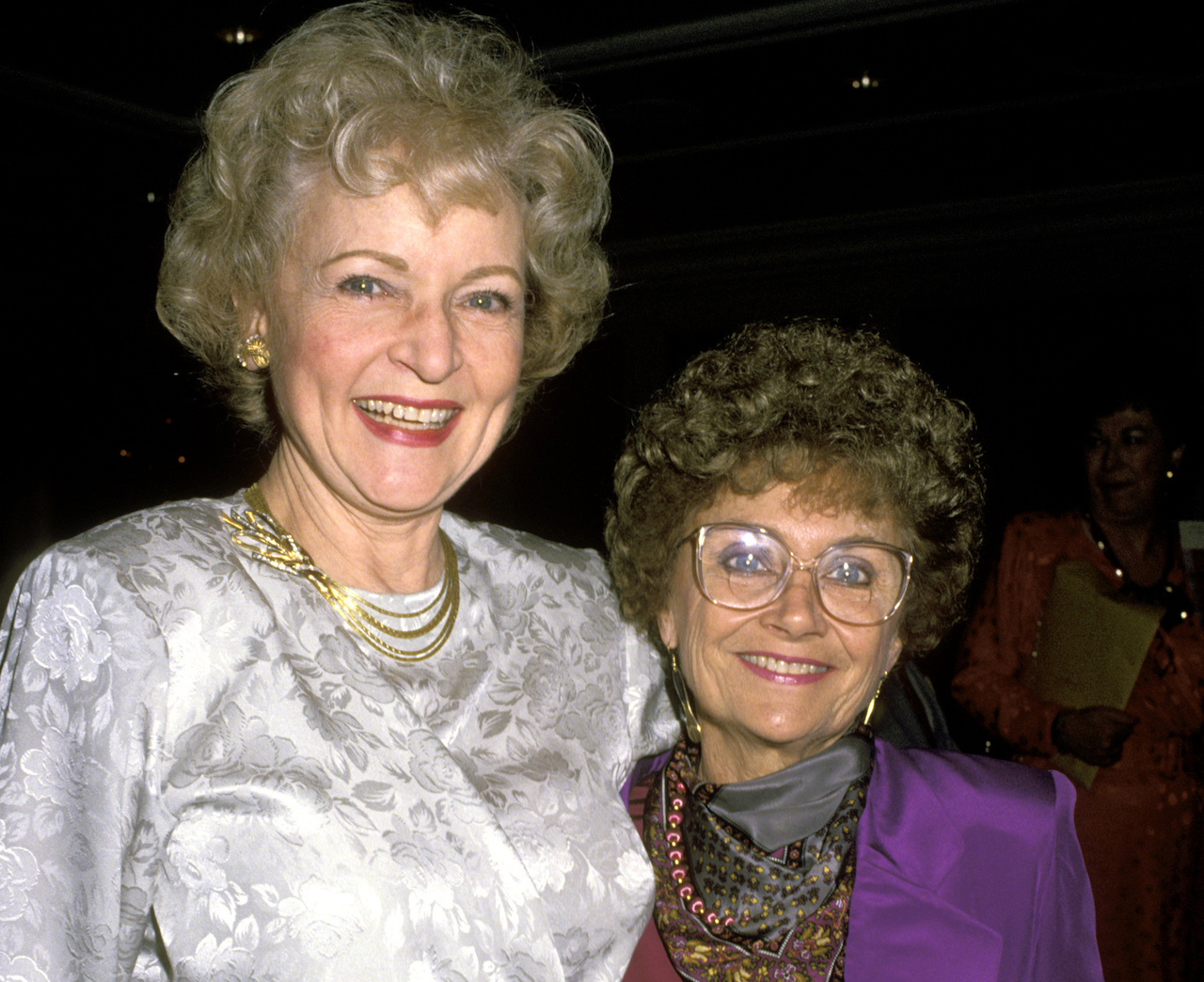 Betty White in a silky, white top, standing with Estelle Getty in purple
