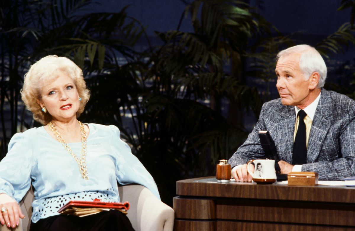 Betty White in a blue top, sitting next to Johnny Carson who is seated at the 'Tonight Show' desk