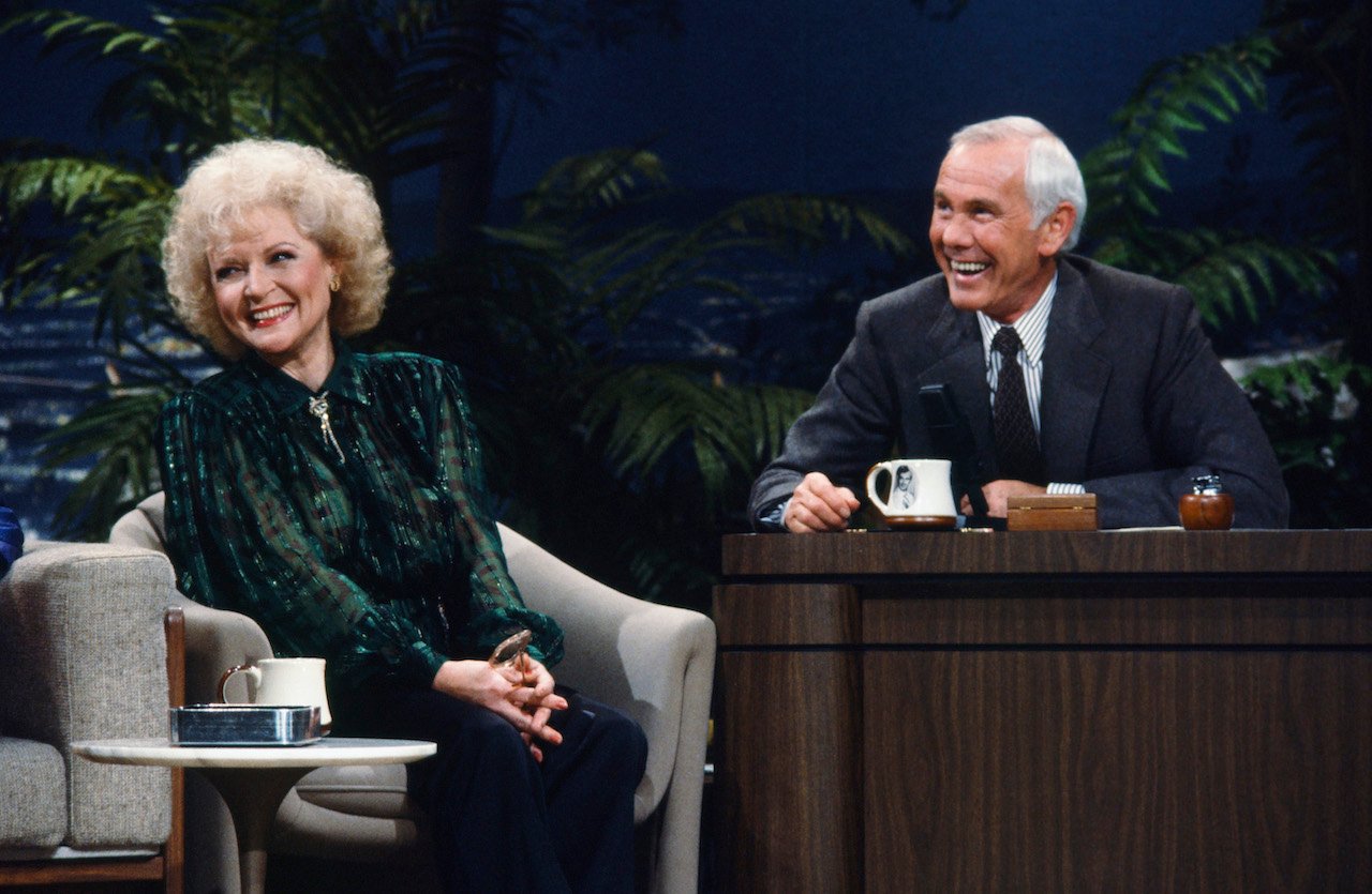 Betty White in a green top, sitting with her hands clasped on her lap and smiling with Johnny Carson, in a suit and also smiling