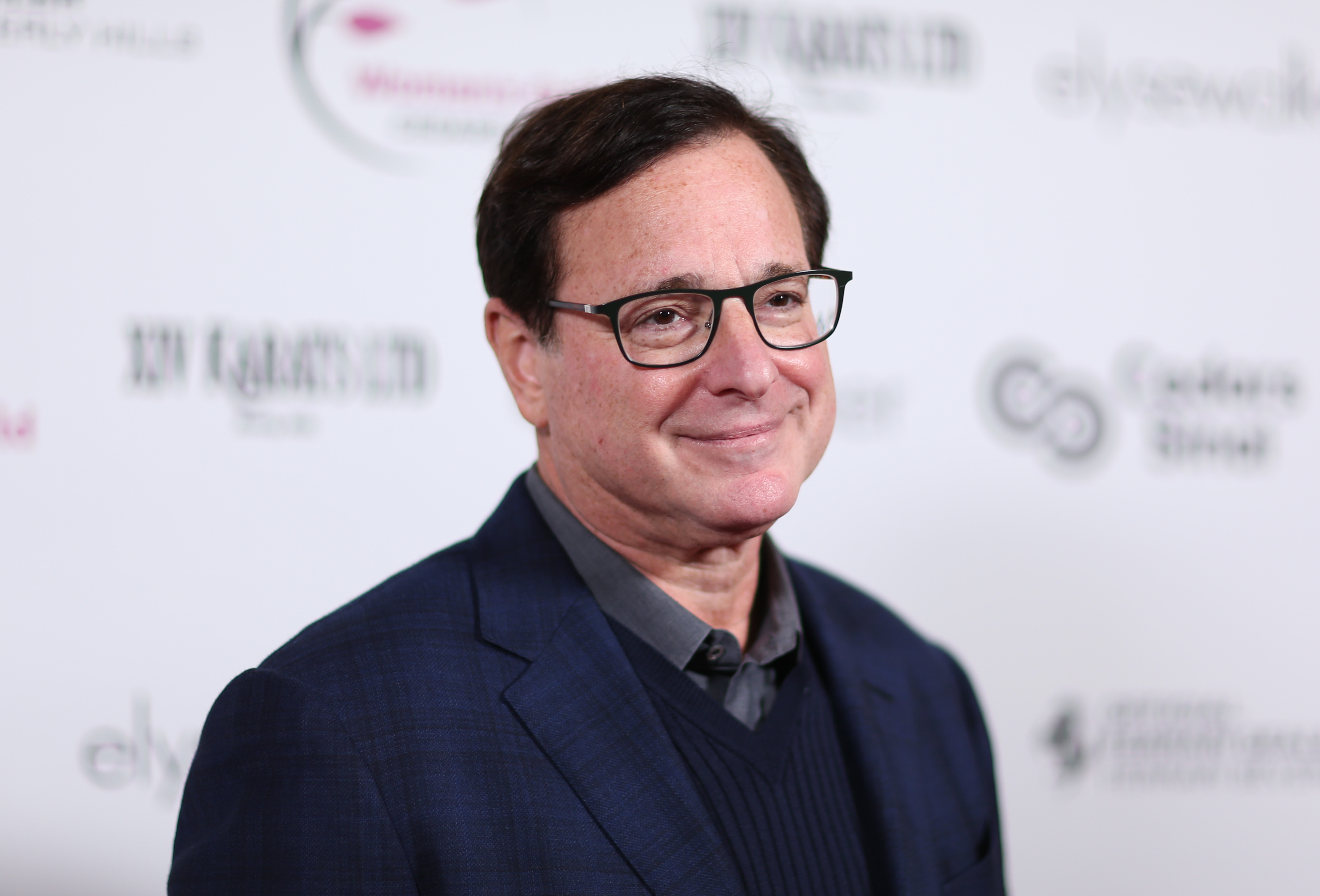 Bob Saget wearing glasses and standing in front of white background