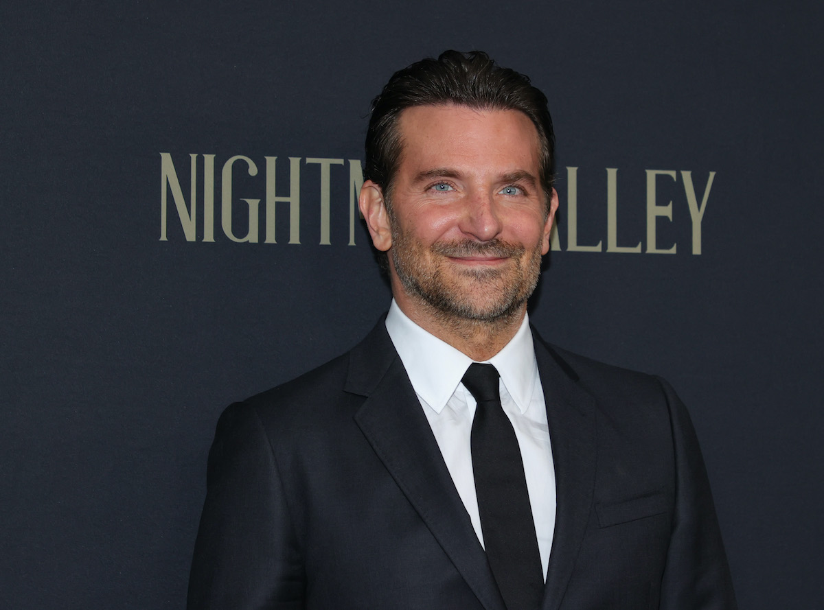 Bradley Cooper wears a dark suit and smiles in front of the ‘Nightmare Alley’ logo