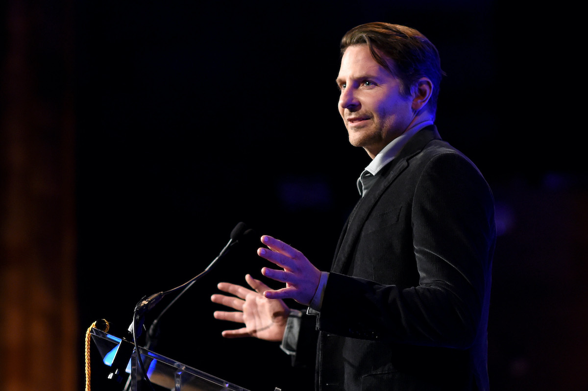 Bradley Cooper wears a dark suit as he stands at a podium speaking with his hands out