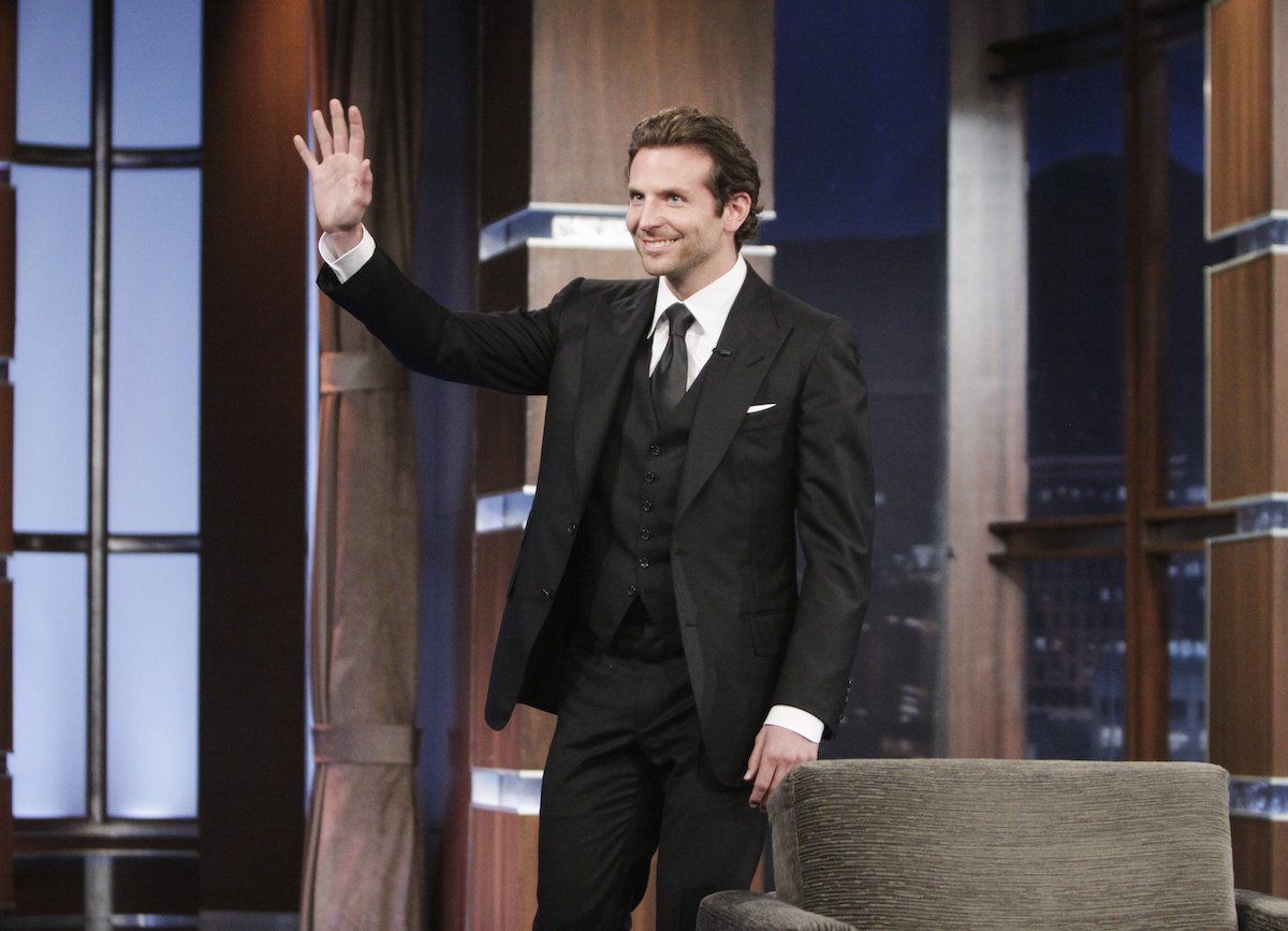 Bradley Cooper wears a dark suit and waves during a talk show appearance
