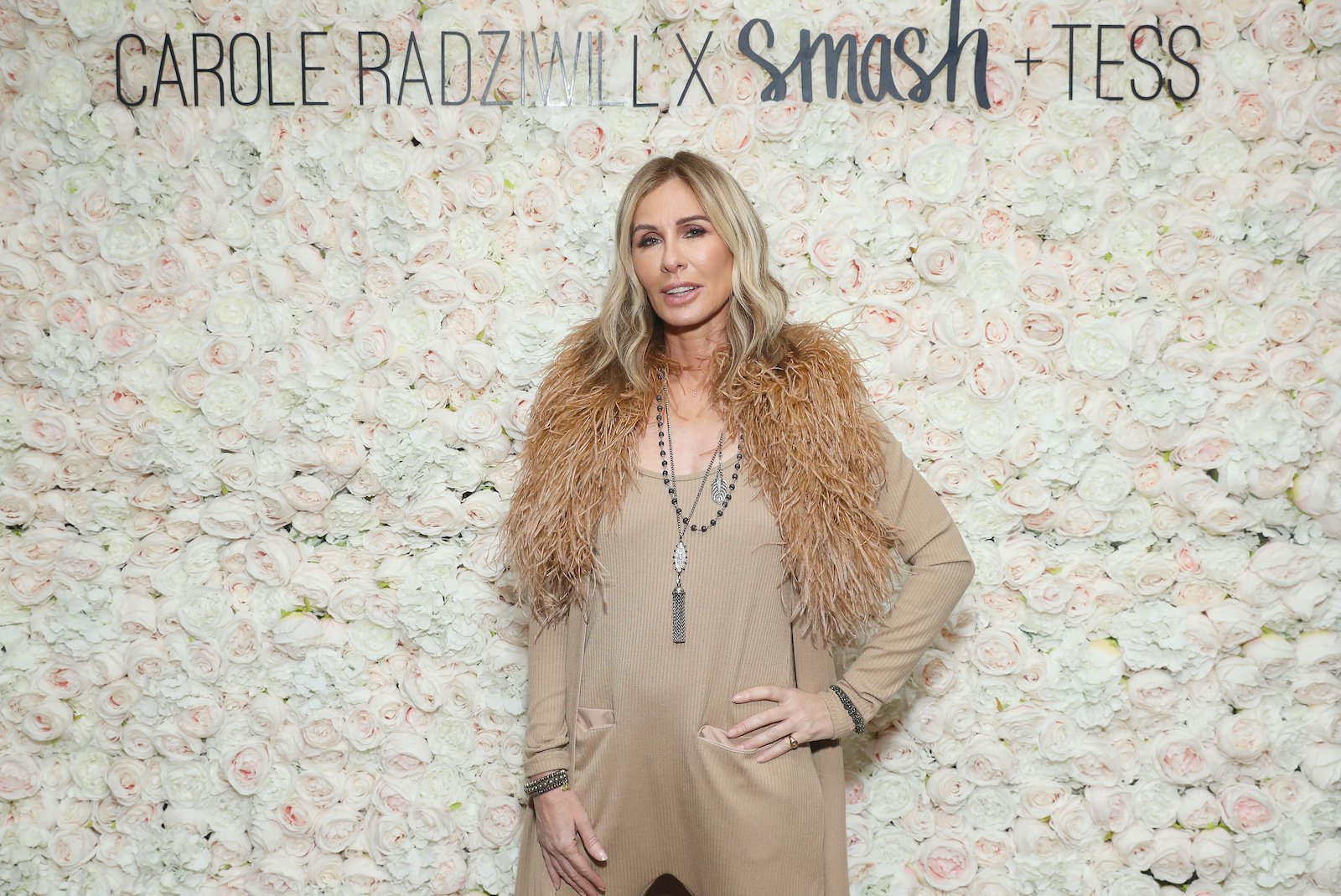 Carole Radziwill wearing a brown and tan outfit poses in front of a flowered backdrop.