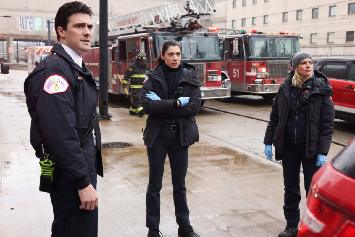 Jimmy Nicholas as Chief Hawkins, Hanako Greensmith as Violet, and Kara Kilmer as Sylvie Brett in Chicago Fire Season 10. The trio stand on the sidewalk in front of two fire trucks.
