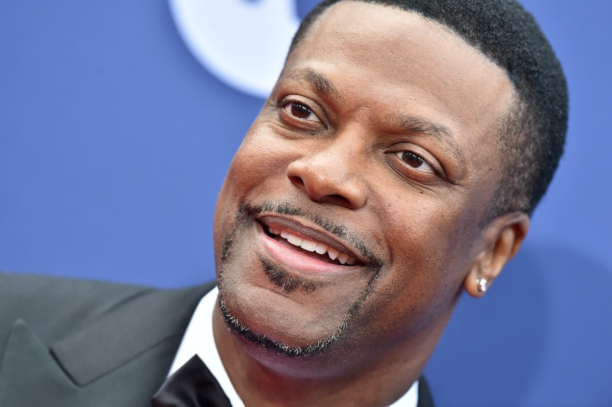 Chris Tucker wears a dark suit and smiles amid a blue background
