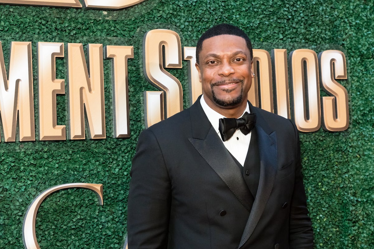 Chris Tucker wears a dark suit and stands in front of a green backdrop on the red carpet