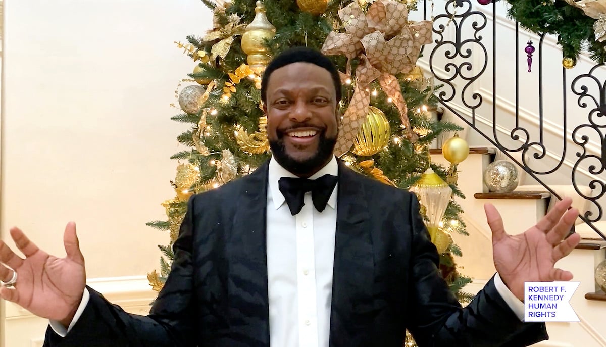 Chris Tucker wears a dark suit as he smiles with arms outstretched in front of a gold-decorated holiday tree