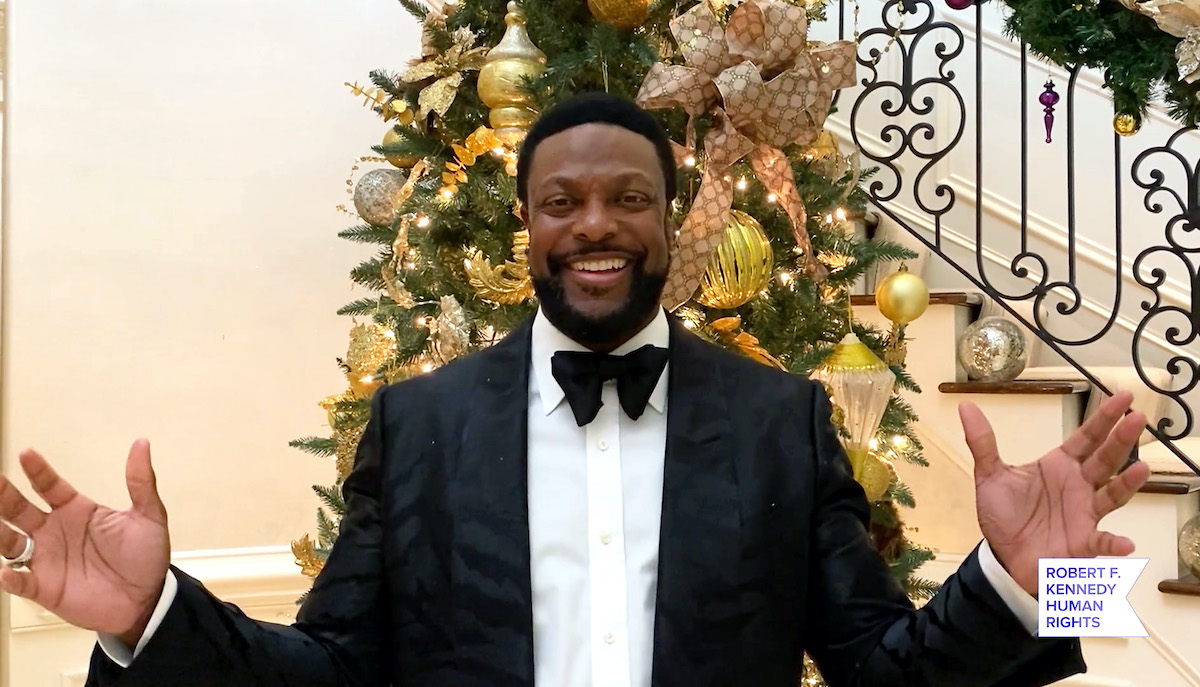 Chris Tucker wears a dark suit as he smiles with arms outstretched in front of a gold-decorated holiday tree