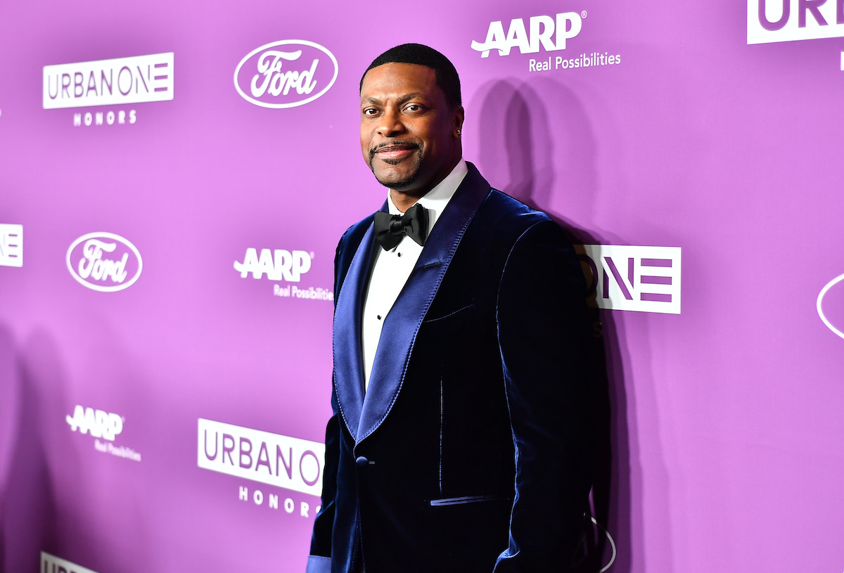 Chris Tucker wears a blue suit and poses in front of a purple backdrop