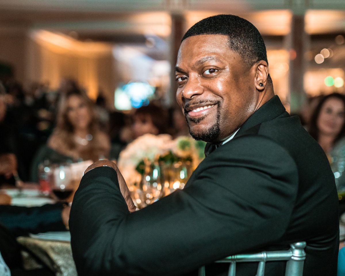 Chris Tucker wears a dark suit and turns to the camera while sitting at a table during an event