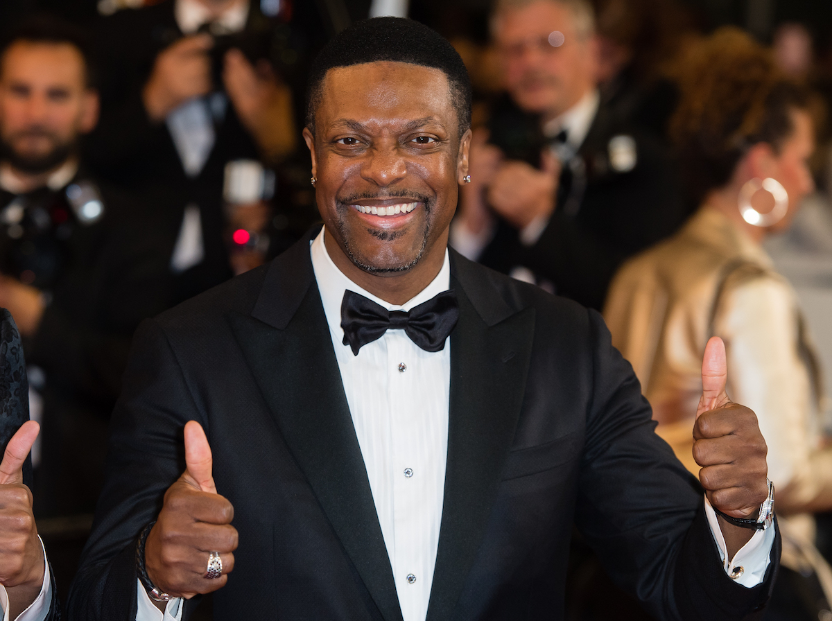 Chris Tucker wears a dark suit and smiles with both thumbs up on his outstretched hands